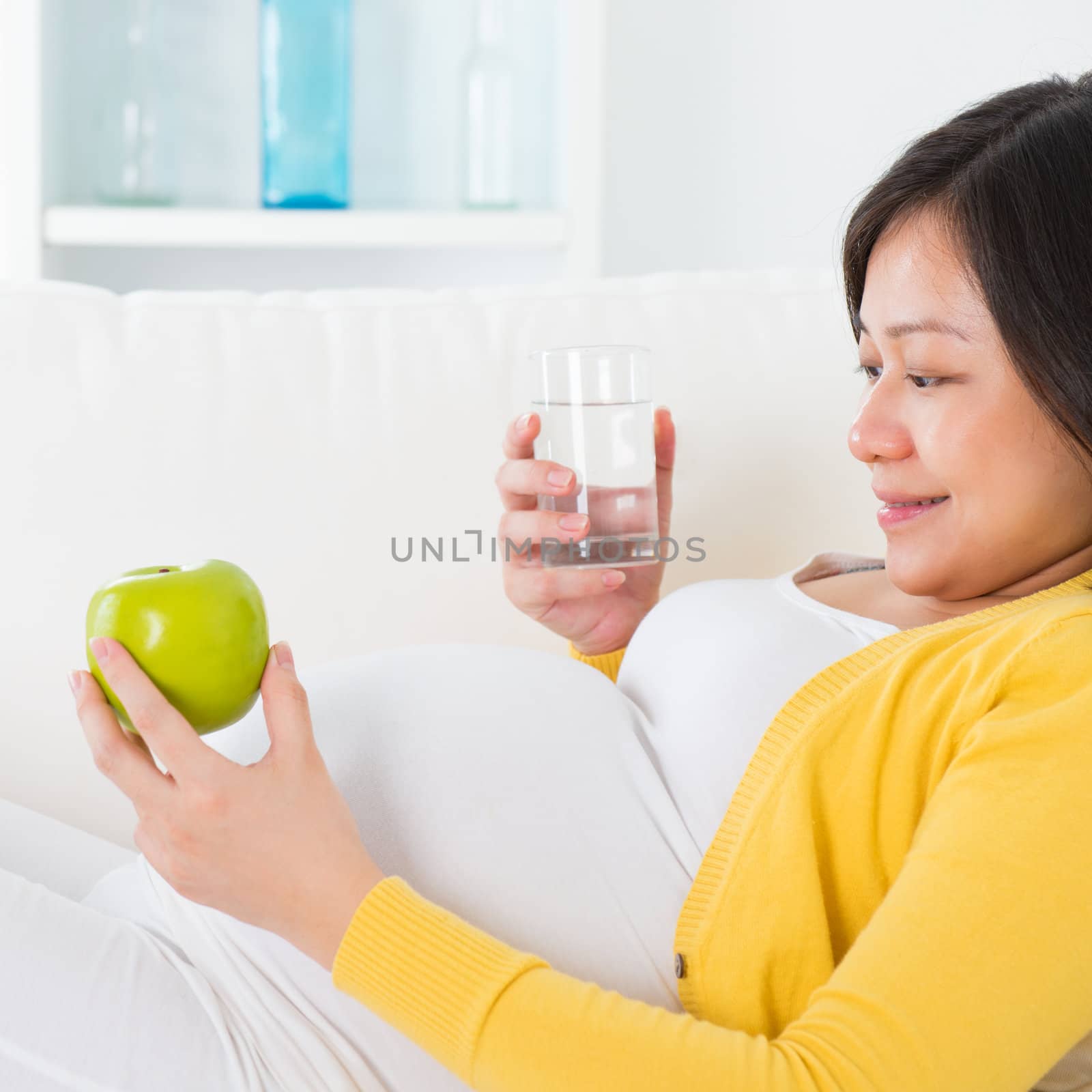 Maternal nutrition. Asian pregnant woman  healthy lifestyle, holding fruit and water glass sitting on sofa at home.