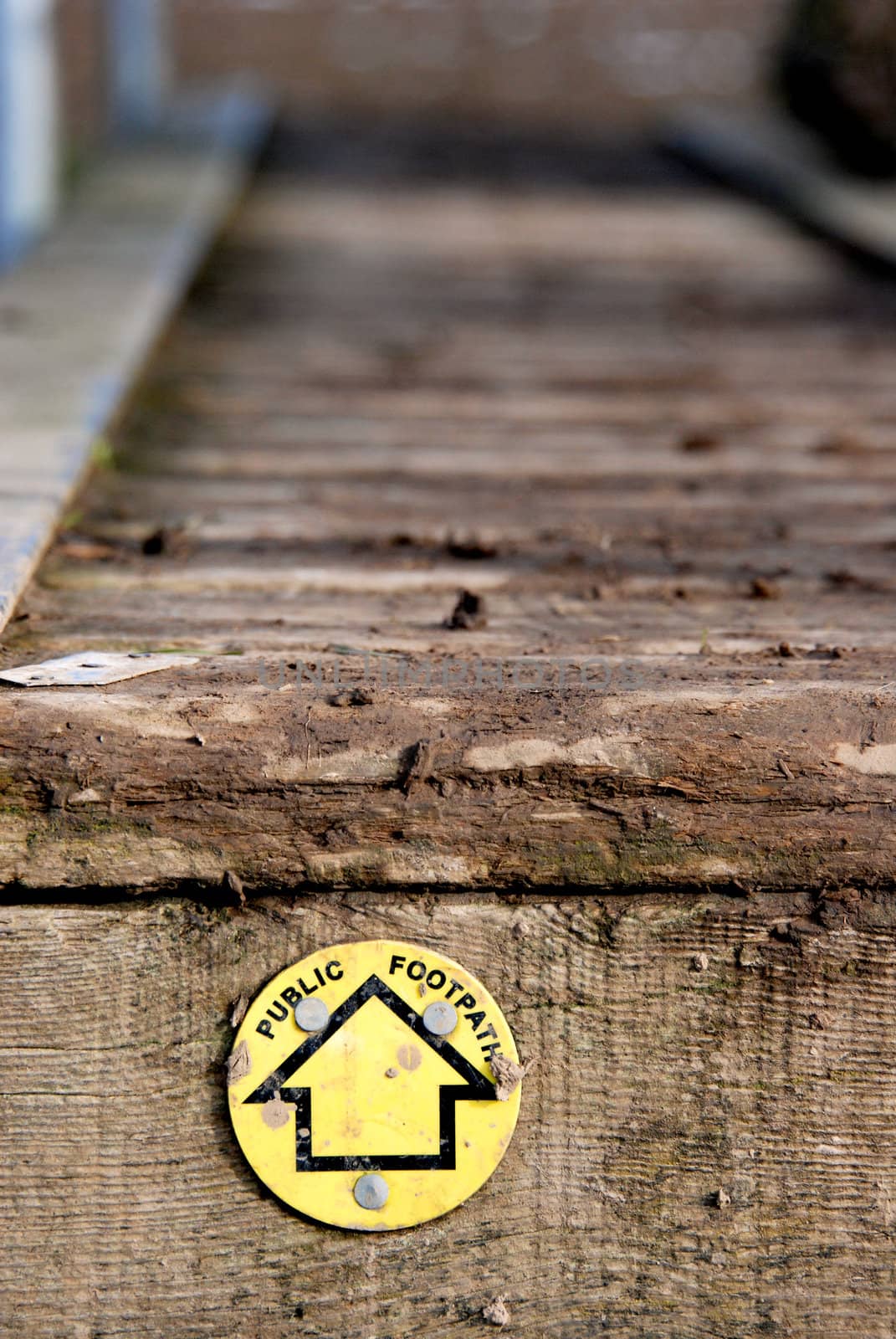 Yellow public footpath sign, leading across a muddy wooden walkway.