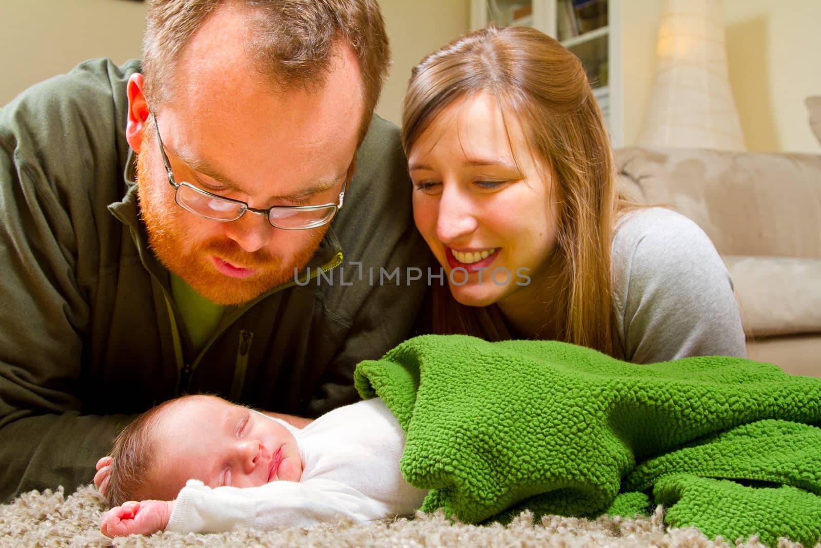 A newborn baby's parents gaze over him while he sleeps. The mother and father look very happy and the baby boy is sleeping peacefully.