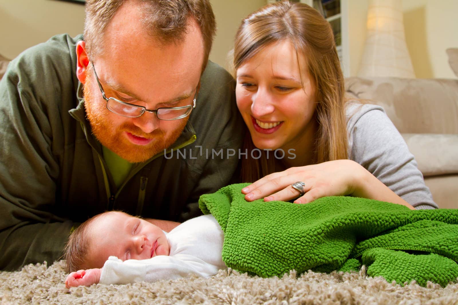 A newborn baby's parents gaze over him while he sleeps. The mother and father look very happy and the baby boy is sleeping peacefully.