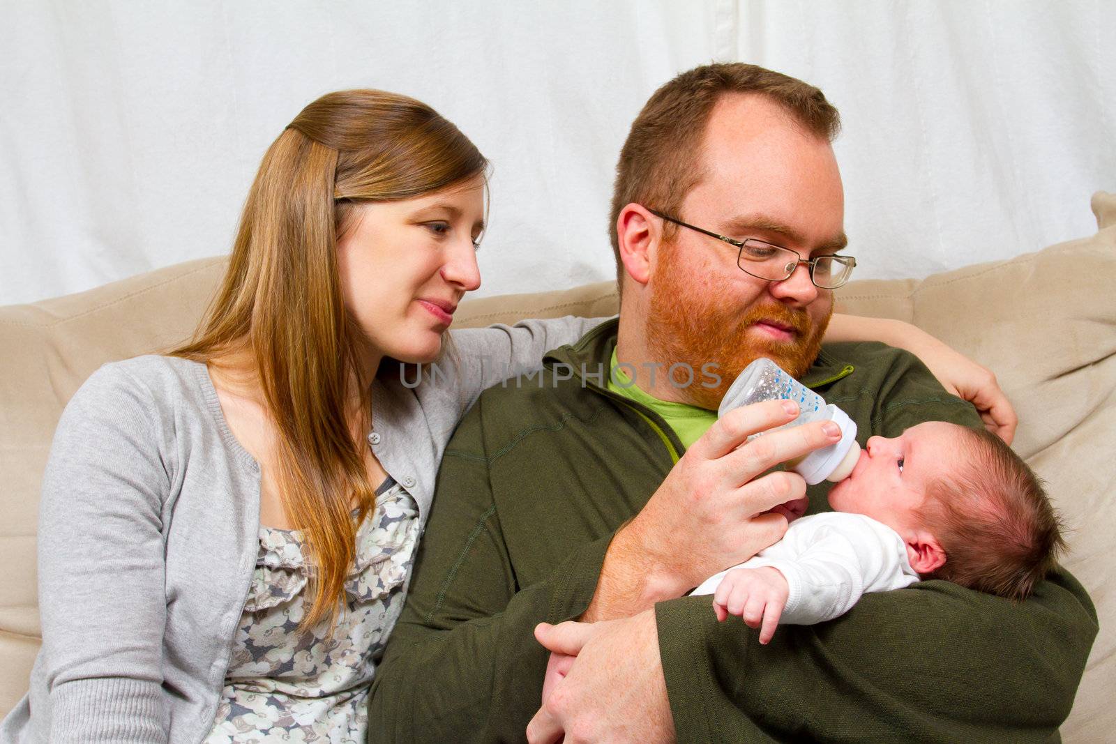 A mother and father hold their newborn baby son in their arms and look towards him showing happiness and caring emotions while bottle feeding the boy.