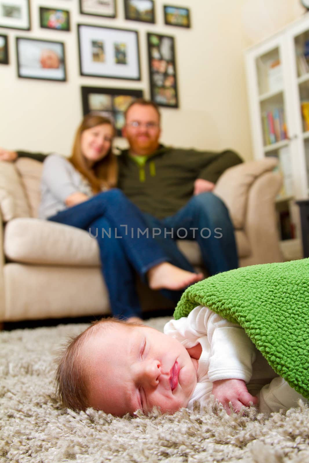 A newborn baby boy sleeps on a comfortable rug while wrapped up in a green blanket with his parents in the background together.