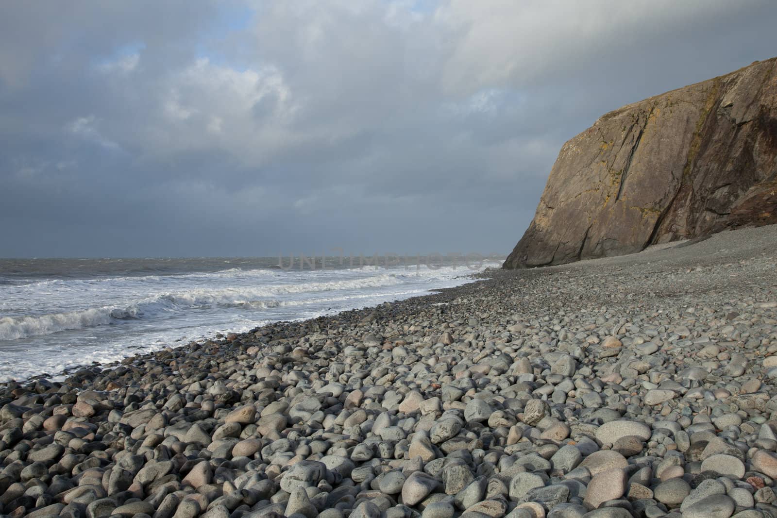 A pebble beach leads to a shale cliff with waves breaking on the shore and a cloudy sky in the distance.