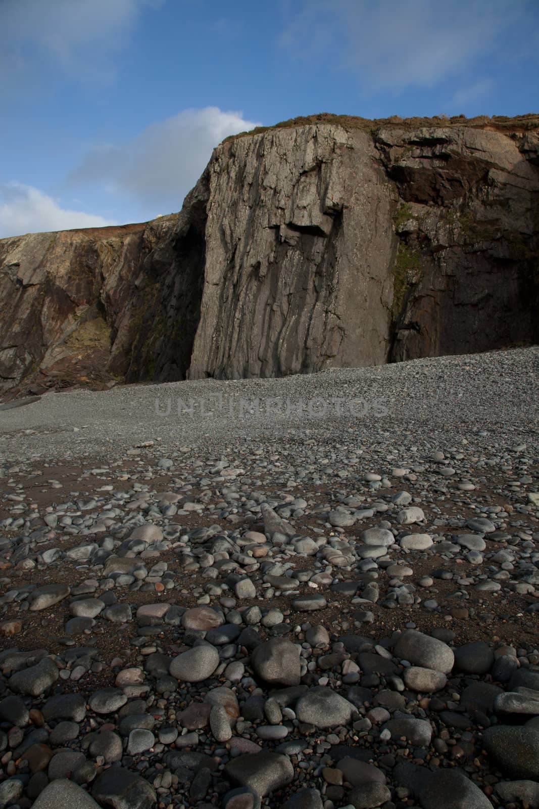 A pebble shore leads to a shale buttress on a cliff with blue sky and cloud in the distance.