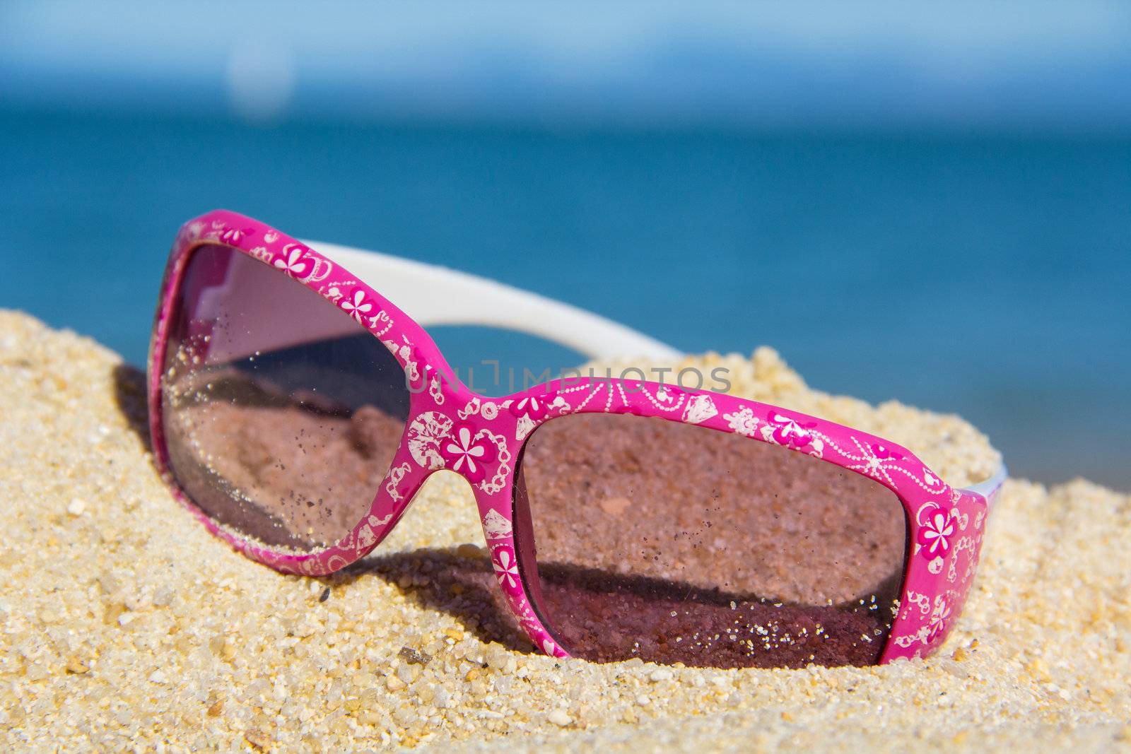 pink kids suglasses with a white floral design on the sand near the sea