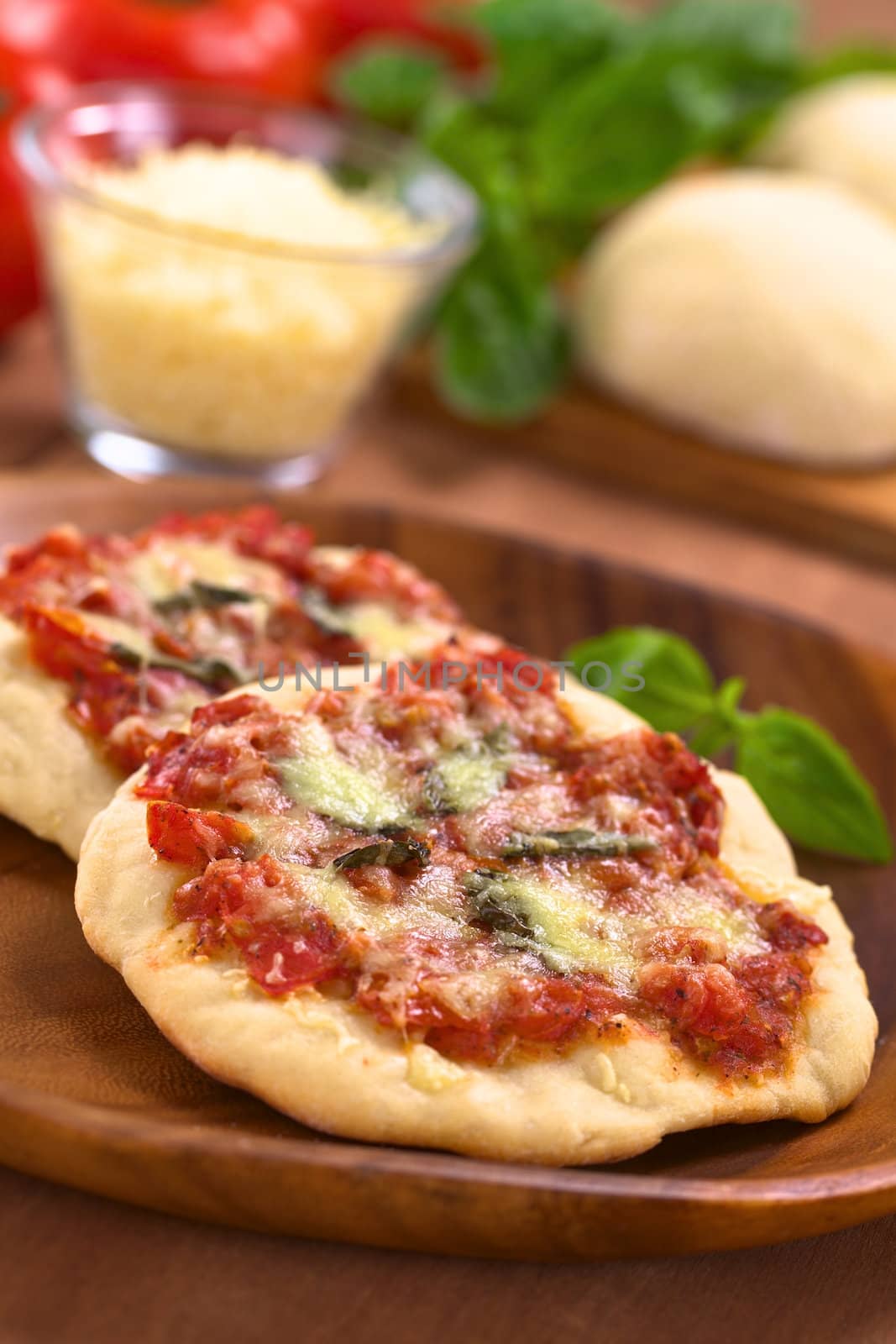 Crispy homemade Pizza Margherita or Pizza Mozzarella (pizza with tomato, basil and cheese) on wooden plate with ingredients in the back (Selective Focus, Focus one third into the first pizza)