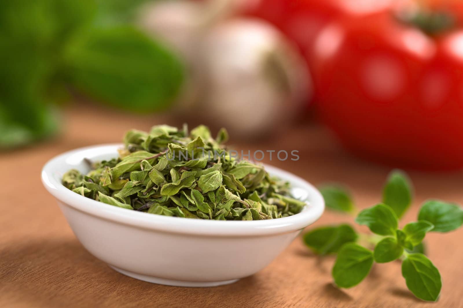 Dried oregano leaves in small bowl with fresh oregano on the side, tomato, garlic and basil in the back (Selective Focus, Focus one third into the dried oregano leaves)