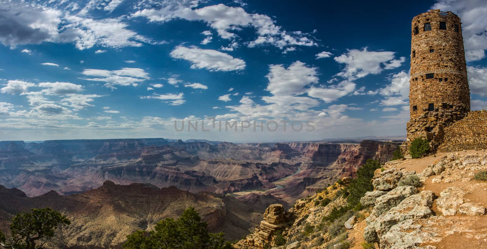 Panorama of the grand canyon with a blue sky and white clouds and the Desert View watchtower to the right of the image