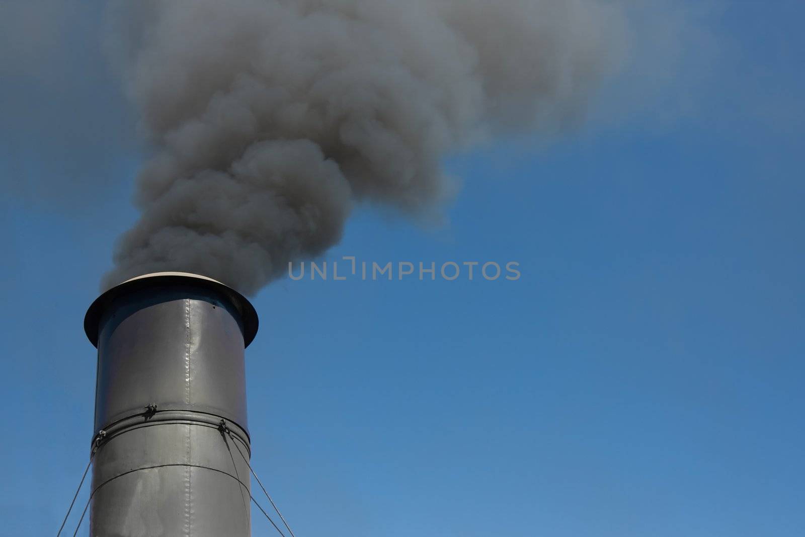 Close up of a metal smoke stack spewing out thick black smoke into a perfect blue sky.