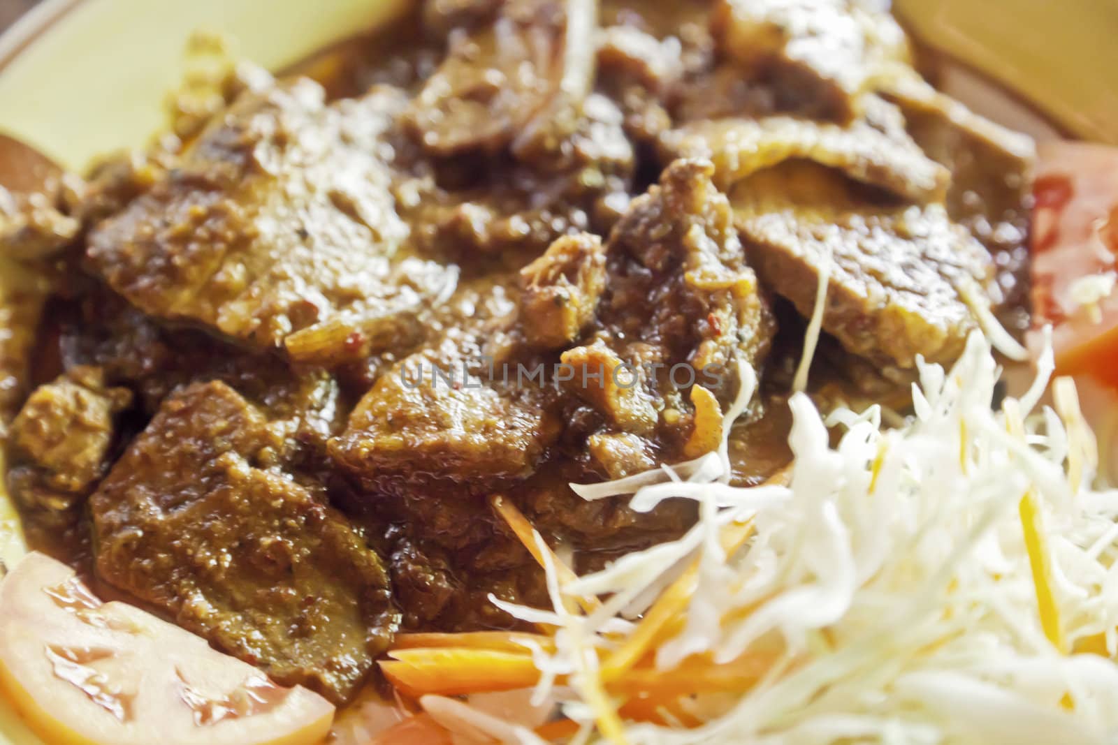Horizontal close up of a plated cooked dish of red meat in thick gravy with shredded cabbage, carrots and sliced tomatoes on a decorated ceramic plate