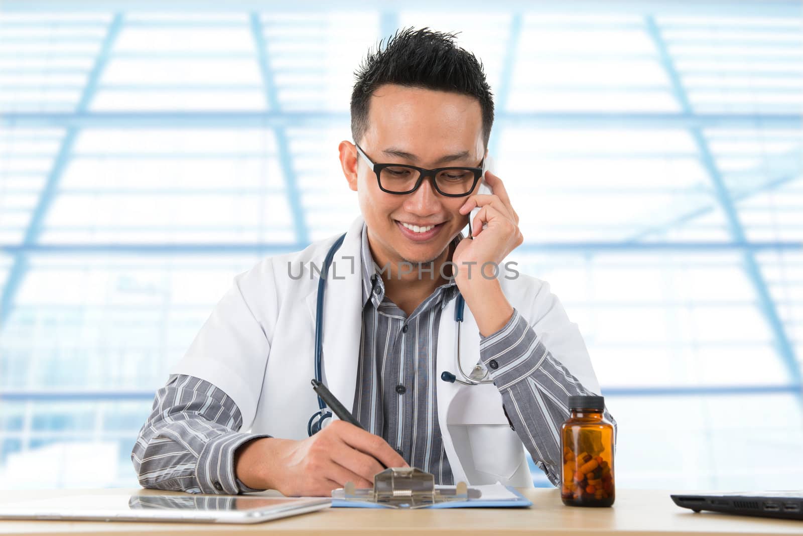 Young Southeast Asian medical doctor writing a note while calling on the phone, sitting in front a desk inside hospital.