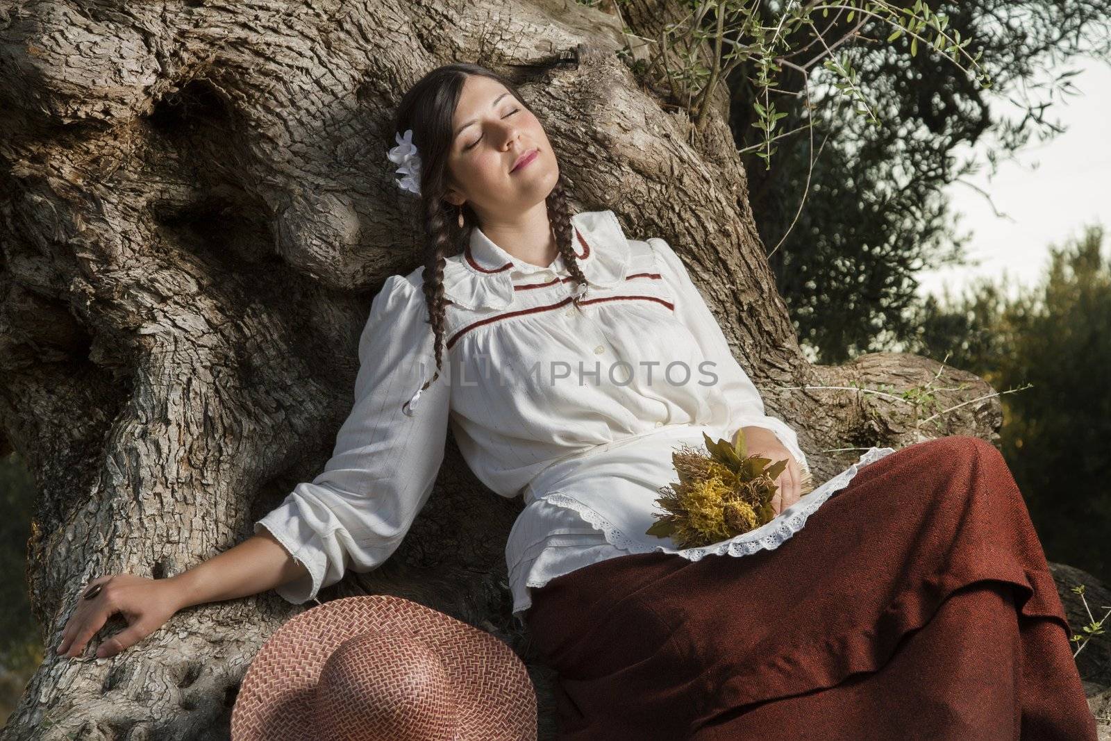 View of a beautiful girl in a classic dress in a countryside set in the shade of a very old olive tree.