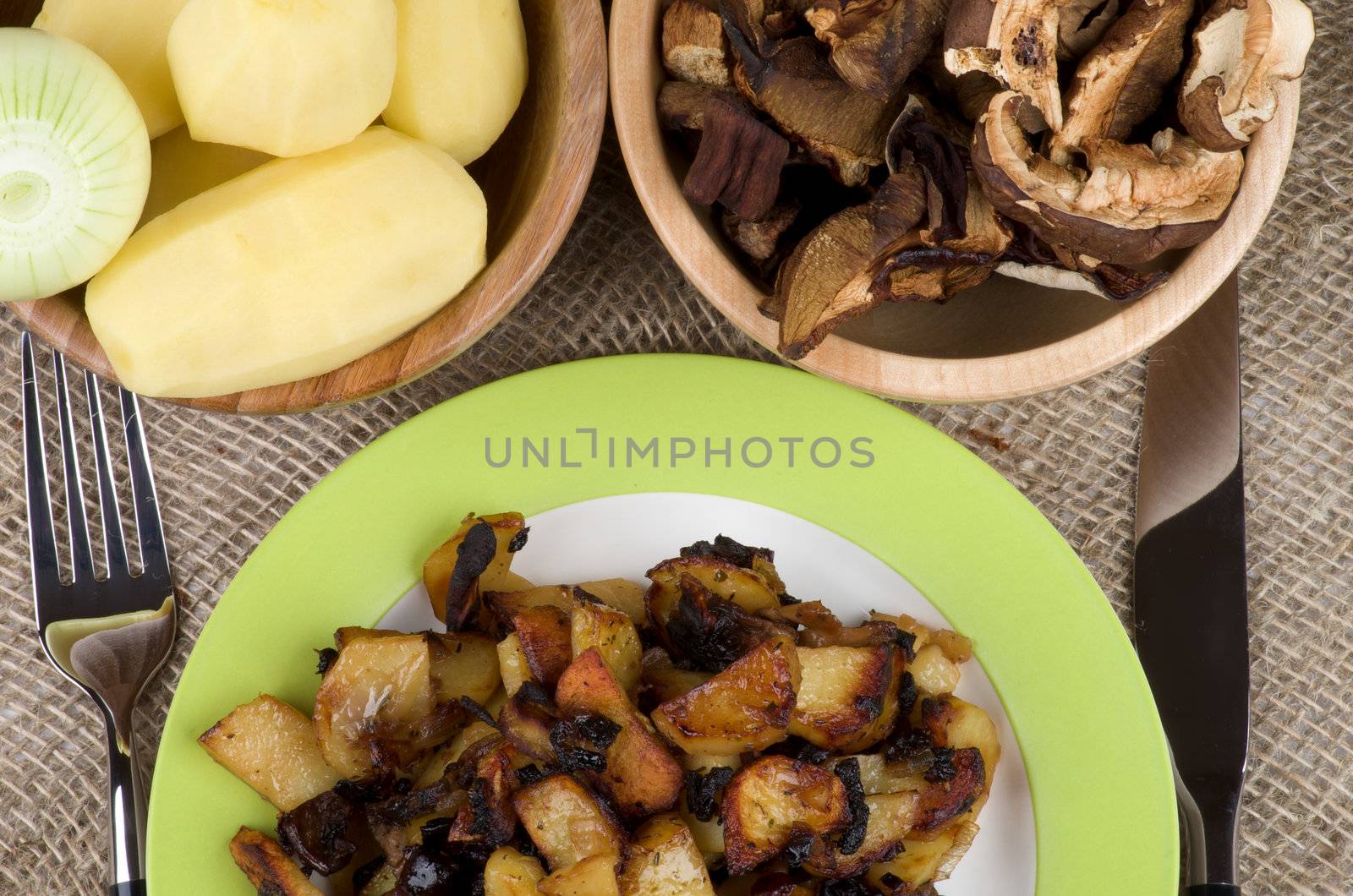 Traditional Roasted Potato with Mushrooms, Onion and Spices. Arrangement of Green Plate with Prepared Potato, Two Wood Bowls with RawPotato, Onion and Slices of Mushrooms and Fork with Knife close up