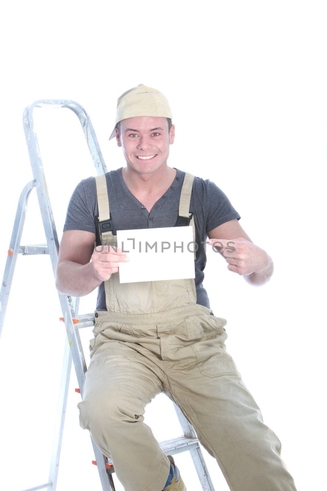 Happy handyman sitting on the rungs of a ladder wearing dungarees pointing to a blank card he is holding in his hand for your advert or text isolated on white