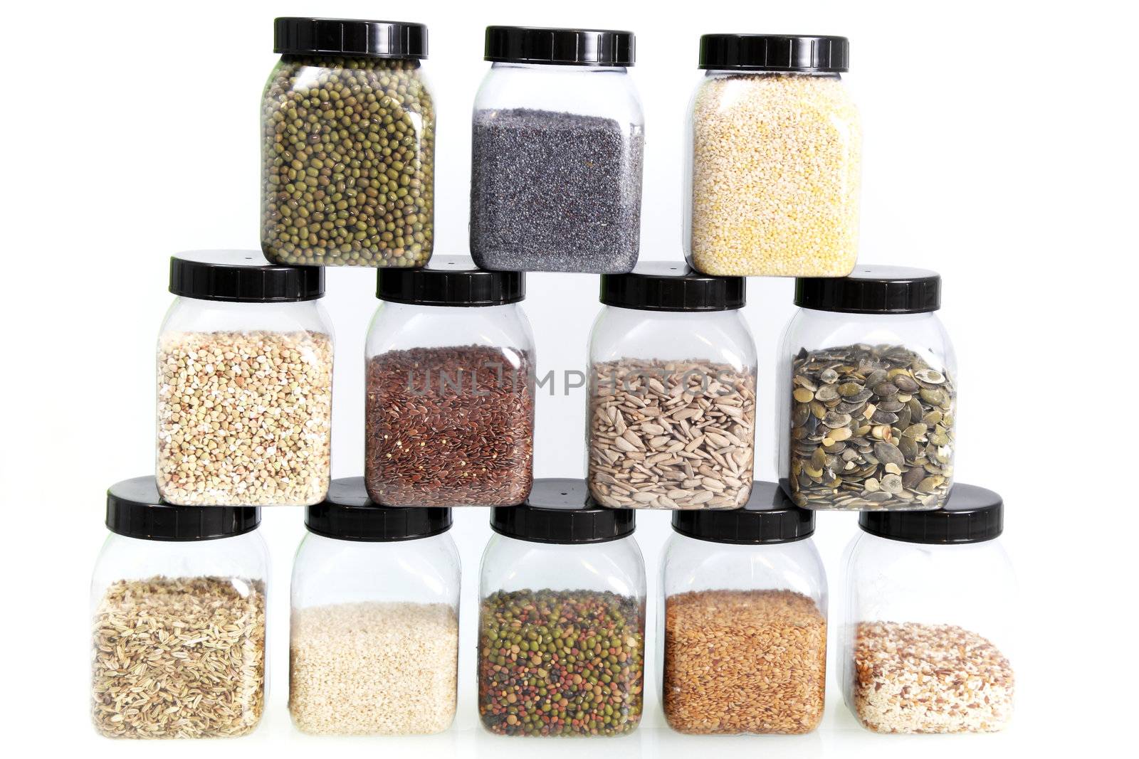 Display of a variety edible seeds in stacked rows of individual glass jars including whole and dehusked sunflower, sesame, poppy, linseed, rice, pulses and legumes isolated on white