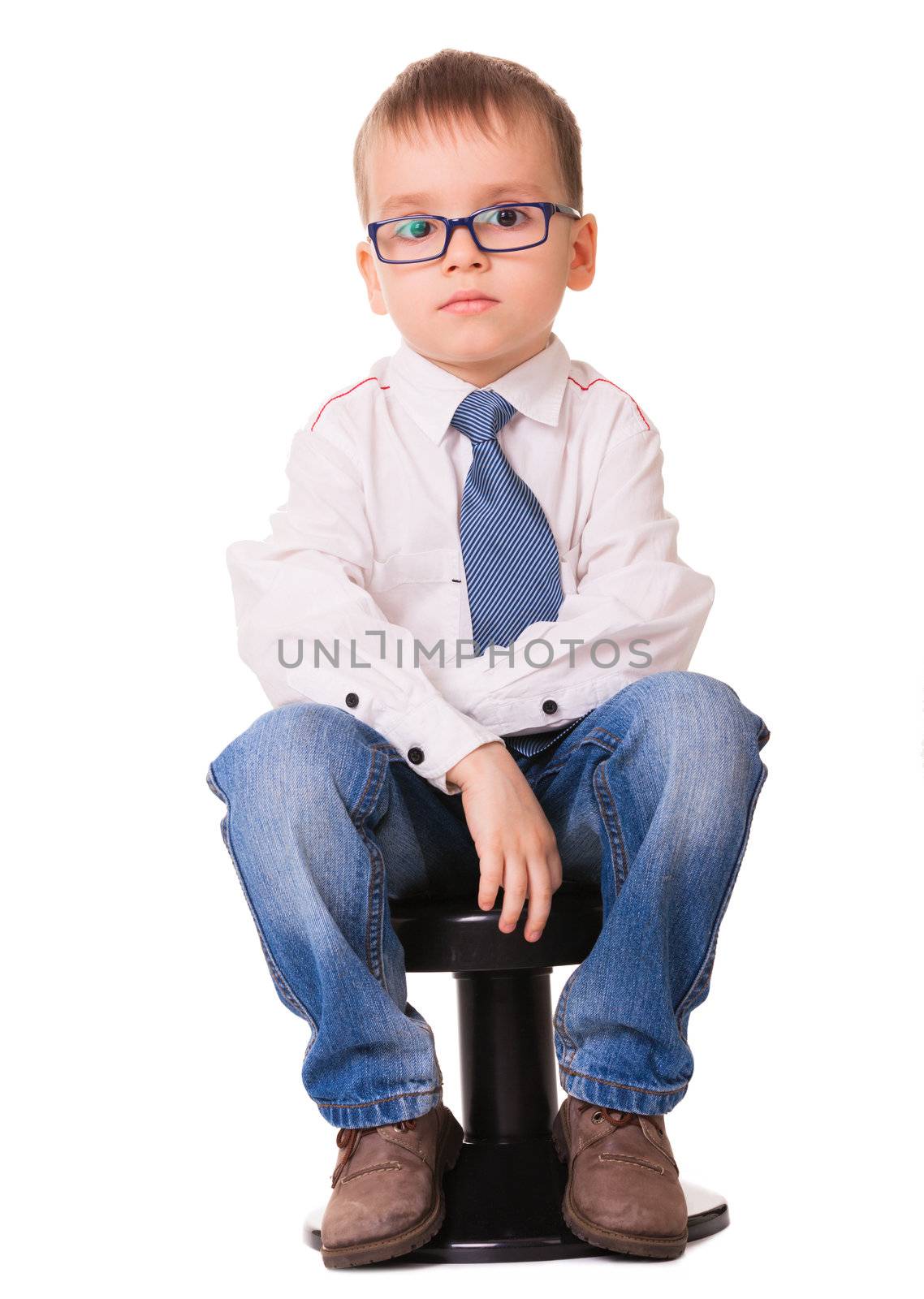  Small sad boy in glasses, shirt and jeans sitting on the black chair. Isolated on white background. 
