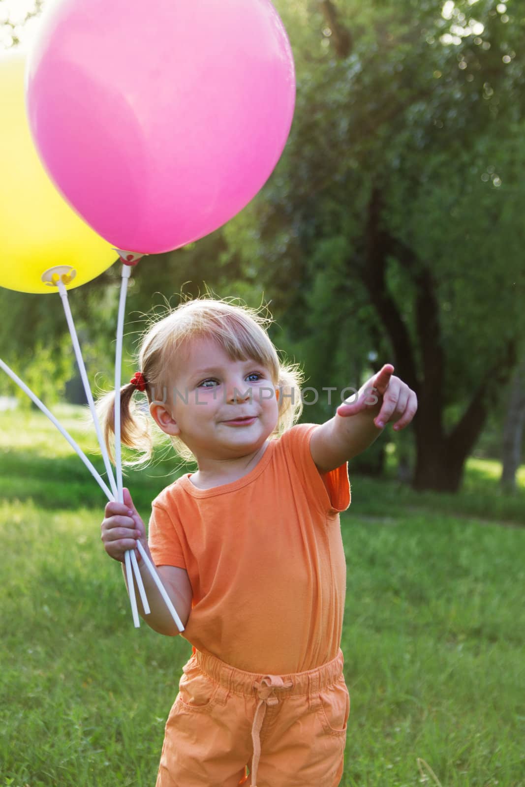 Happy baby girl with balloons among meadow pointing at smth