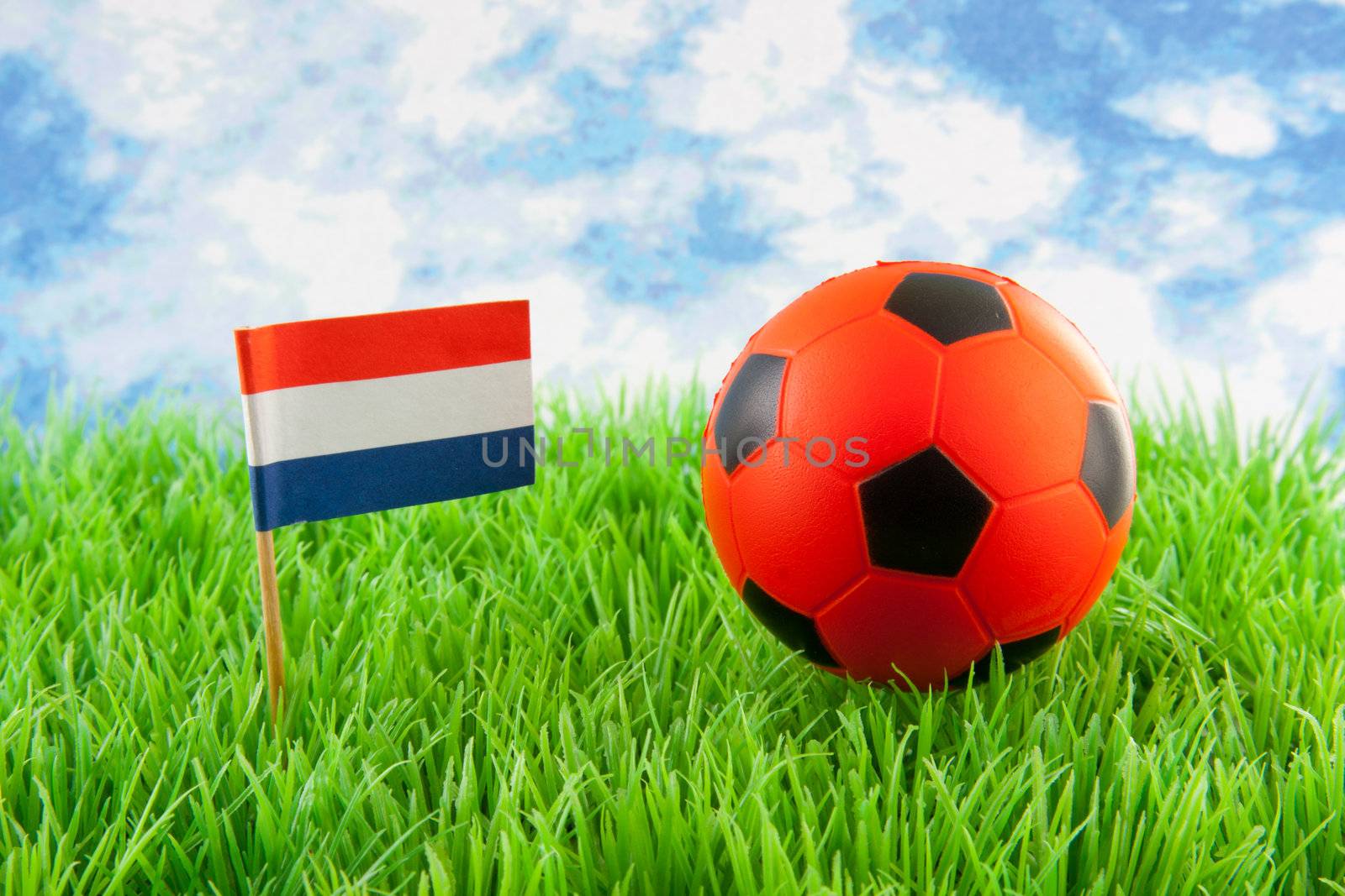 Orange ball and Dutch flag on soccer field against paper blue cloudy sky 
