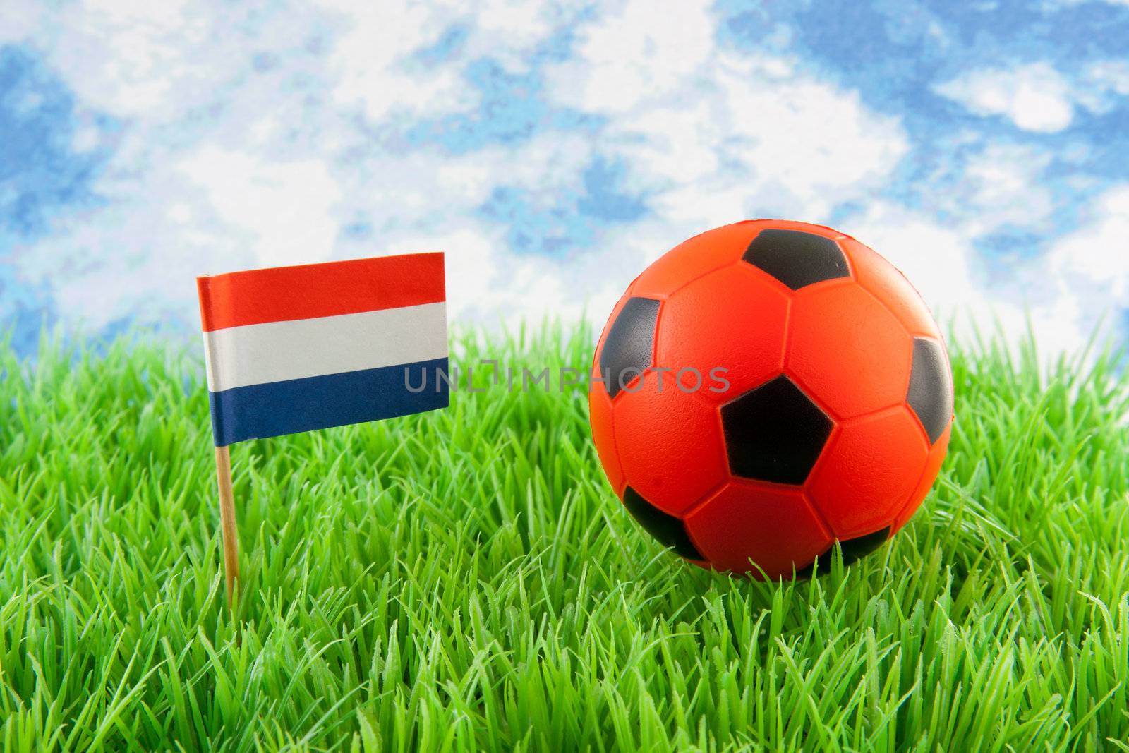 Orange ball and Dutch flag on soccer field against paper blue cloudy sky 