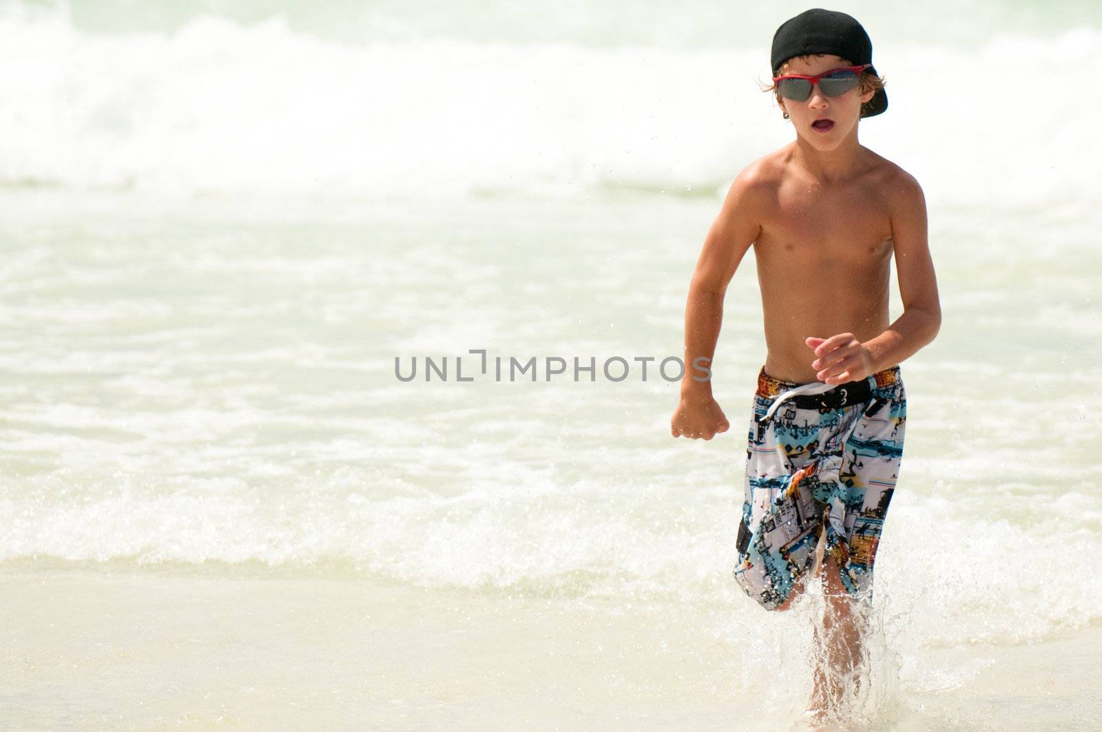 Young boy running through the ocean water wearing sunglasses and backwards hat.