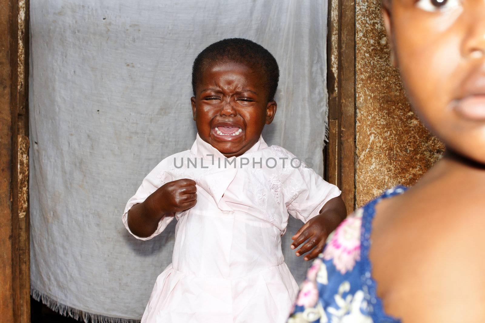 Caught on the fly: little black African baby girl, crying loudly, with bigger sister in the foreground; focus on crying toddler; candid, natural and unstaged portraits.