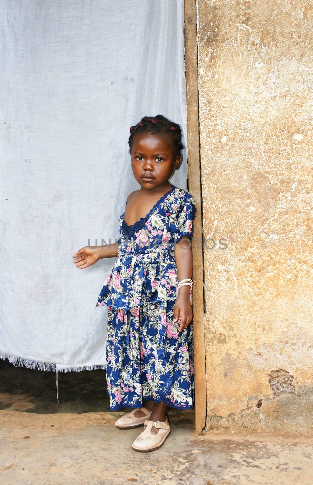 Cute but serious little black African girl in pink sunday dress next to her home door made of fabric; third world or developing country concept.