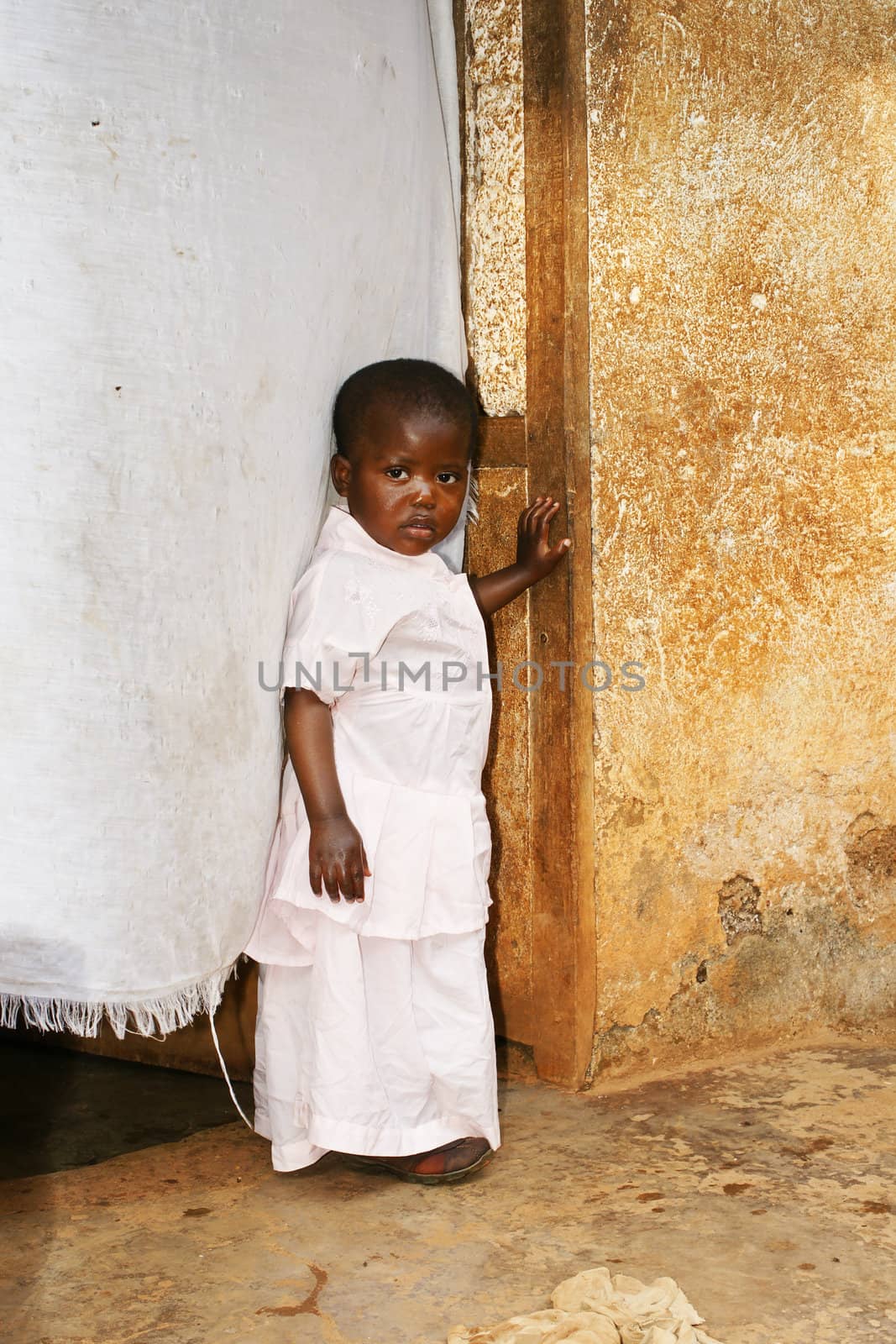 Cute but sad little black African girl in pink sunday dress next to her home door made of fabric; third world or developing country concept.