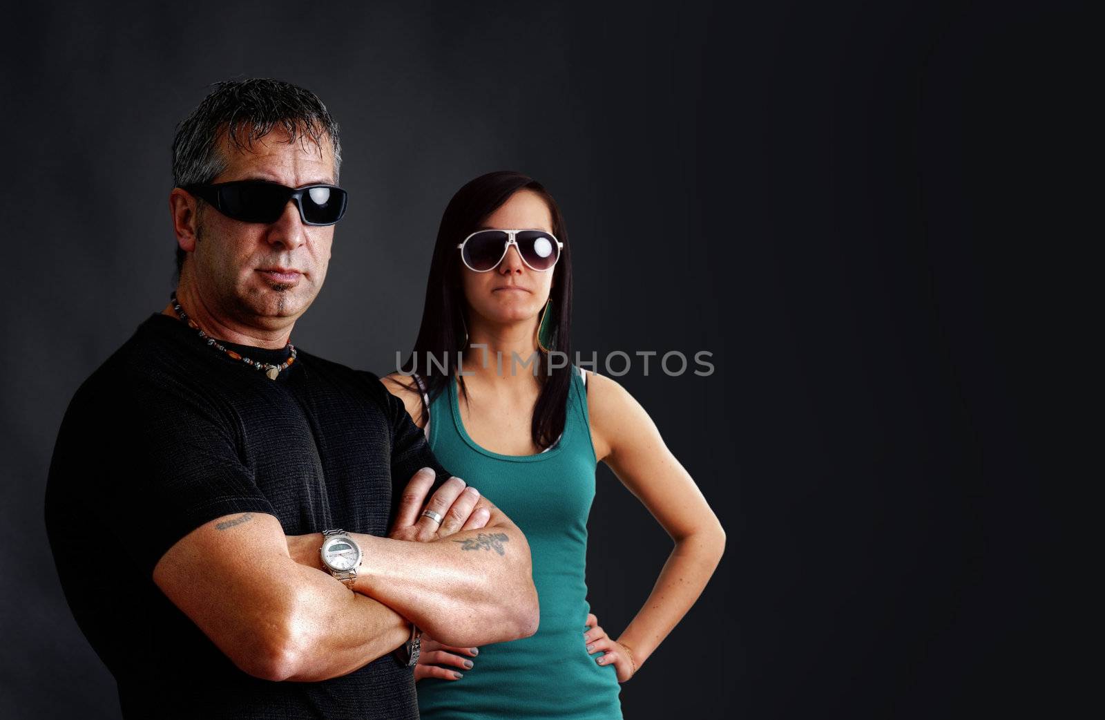 Biker couple: tough guy with tatoos and black sunglasses, arms crossed, looking at camera with pretty young brunette woman, studio shot over black.