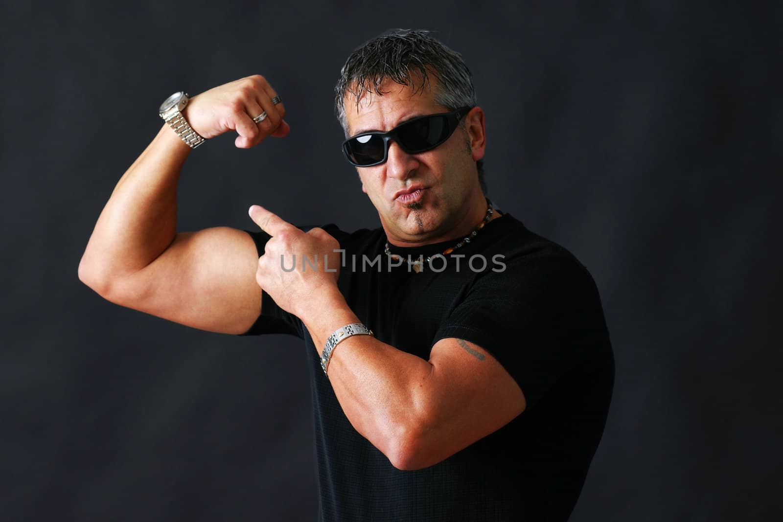 Tough guy with dark glasses, could be criminal, showing his big bicep muscle has an intimidation tactik, studio shot over black.
