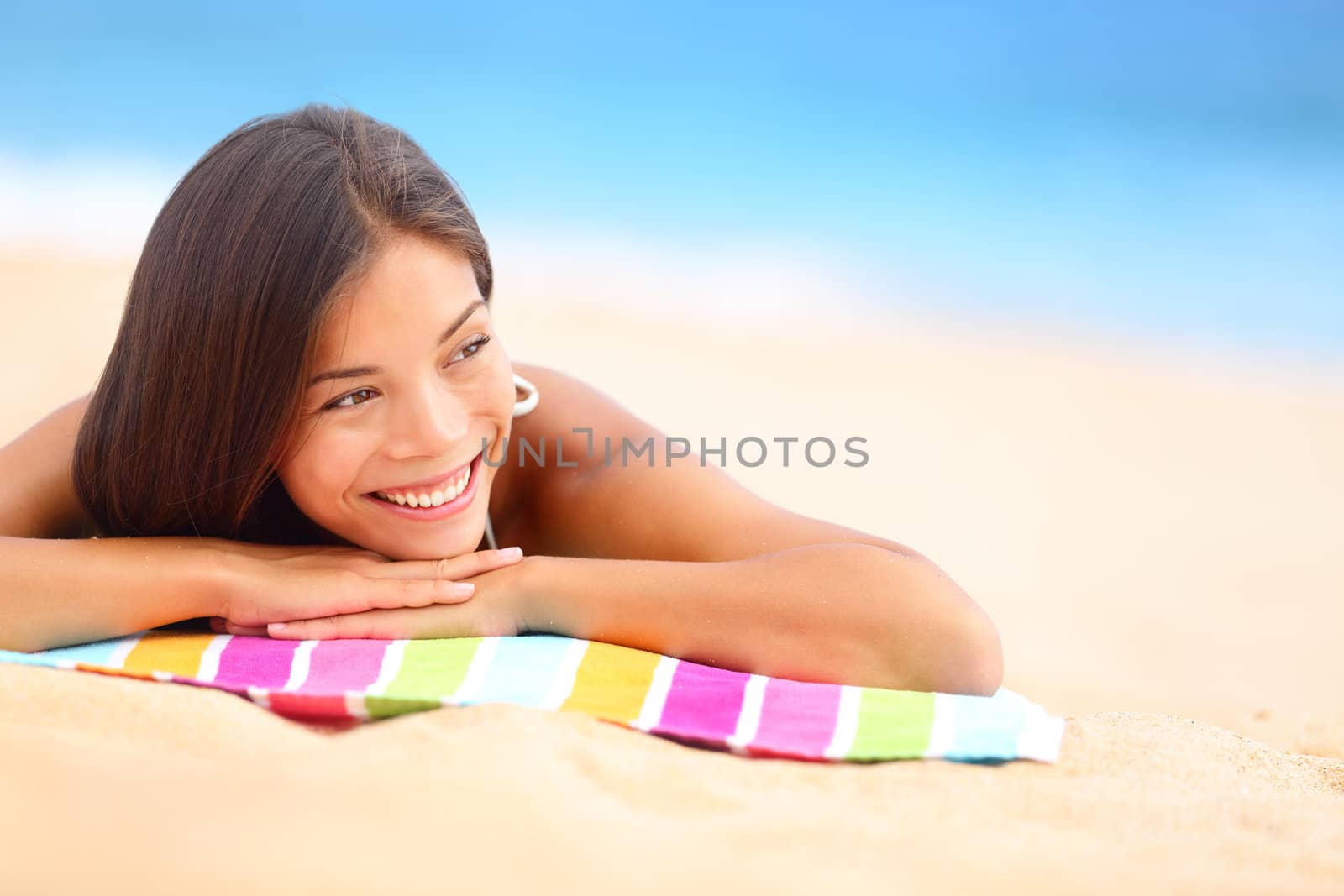 Relaxing beach woman smiling happy looking to the side. Beautiful girl sunbathing under summer sun lying in sand on beach with blue water. Mixed race Asian Chinese / Caucasian female model.