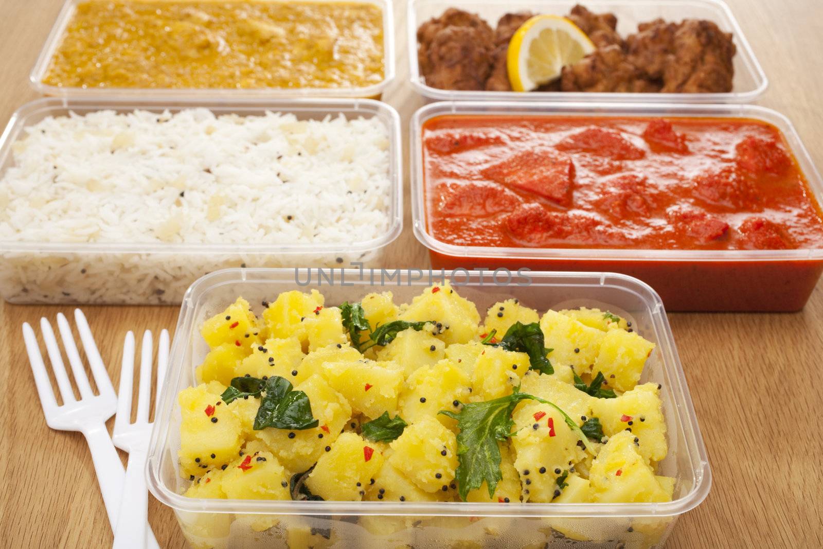 A selection of Indian takeaway food in plastic containers on a wooden table. Aloo saag (potato spinach curry), chicken tikka masala, chicken bhoon or  bhuna, basmati rice and onion bhaji or pakora.