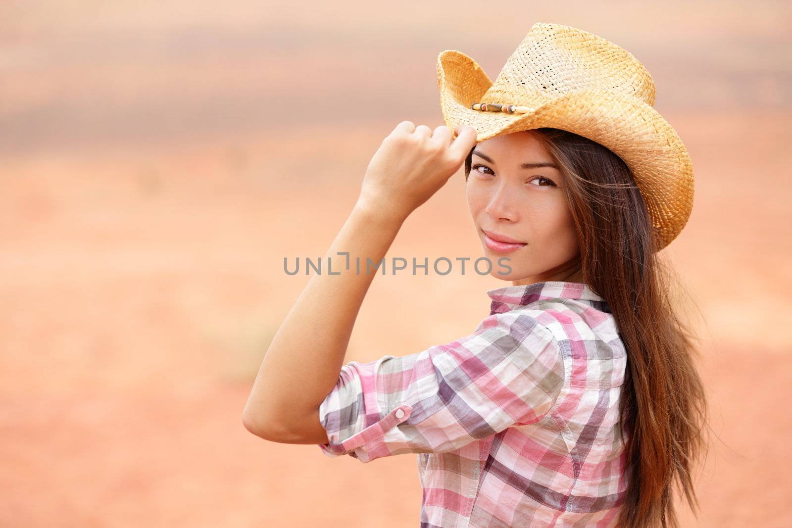 Young american cowgirl woman portrait outdoors. Beautiful natural woman saying hello looking at camera touching cowboy hat. Multicultural Caucasian / Asian girl in her twenties outdoor in nature.