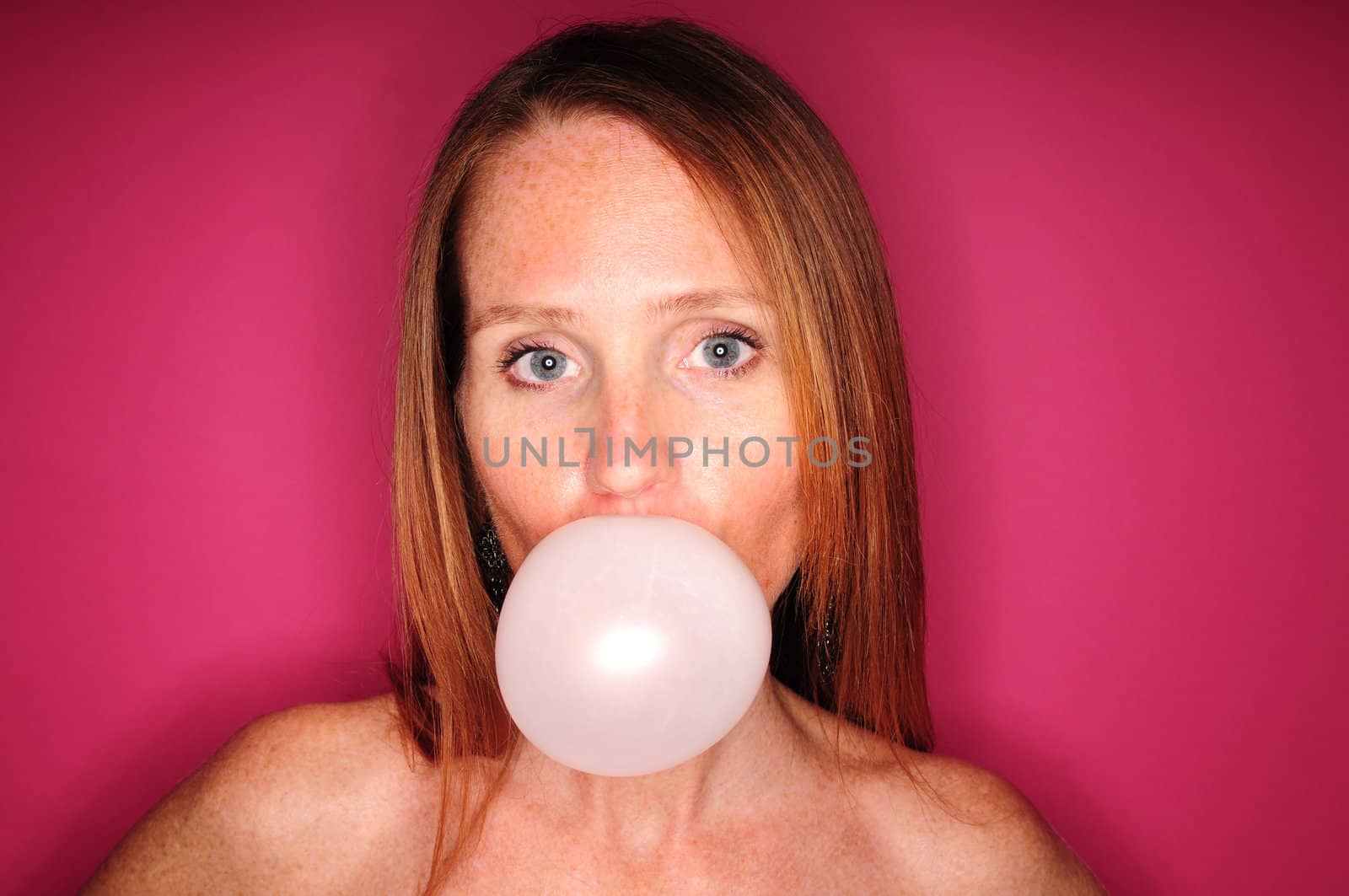 Young woman with red hair and blue eyes blowing a bubble with chewing gum on a pink background