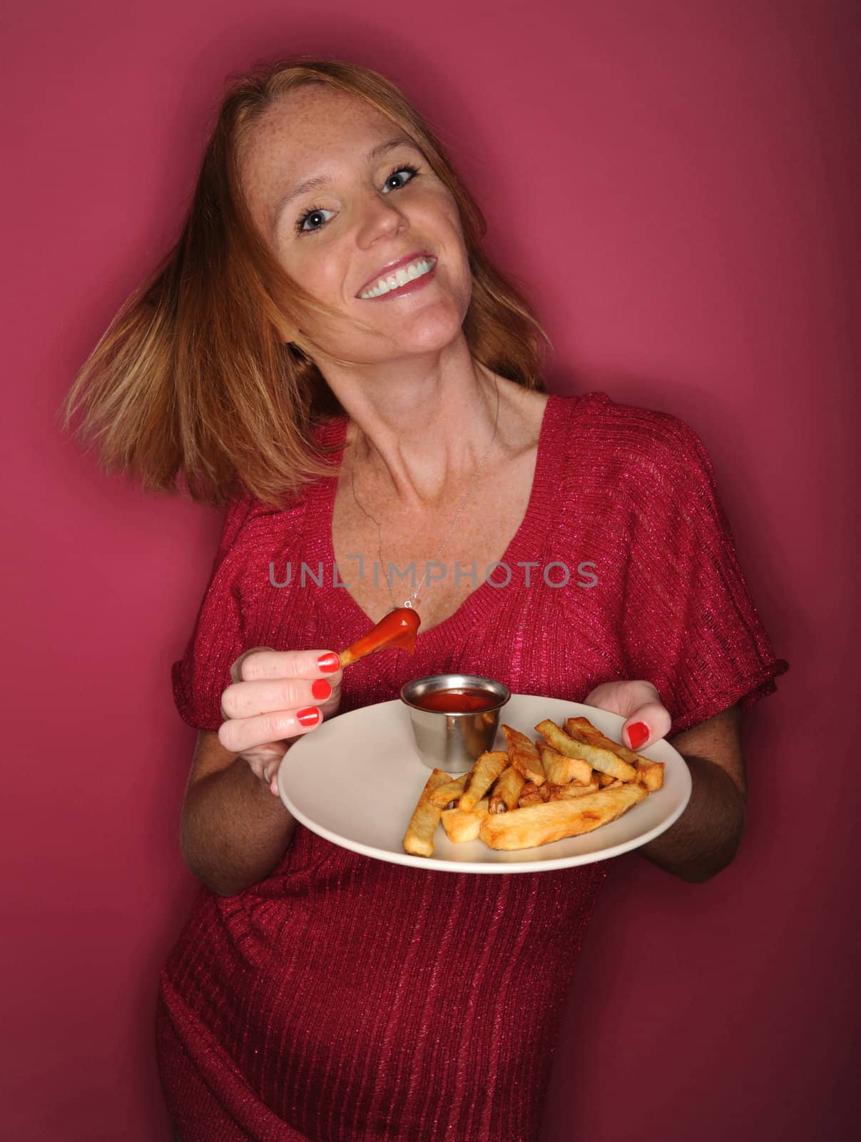 A pretty redhead smiling and enjoying French fries on a pink background