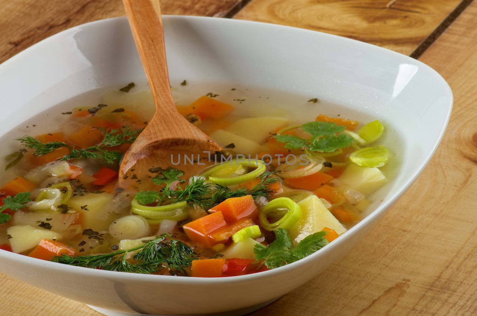 White Bowl of Vegetable Soup with Potato, Carrot, Bell Pepper, Leek, Parsley and Dill with Wooden Spoon closeup on Wooden background