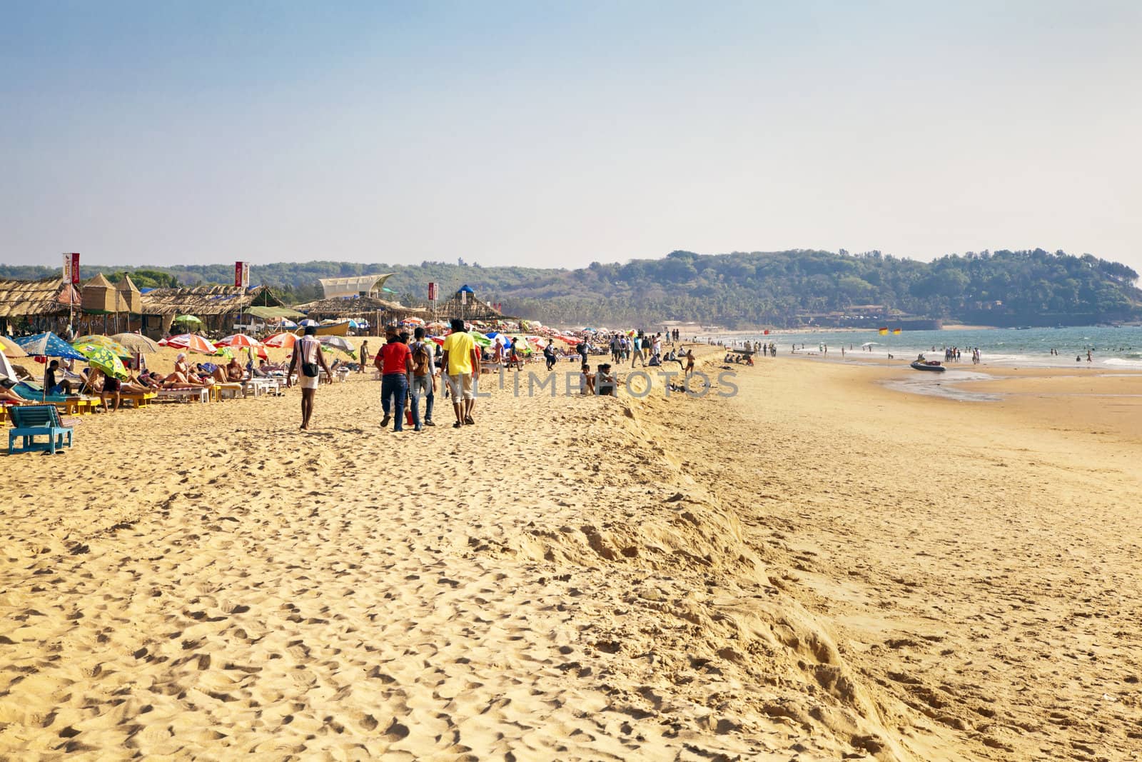 Horizontal capture of a group of young men strolling on a busy sandy beach full of holiday makers on a sunny afternoon. Location of this landscape was Goa, India