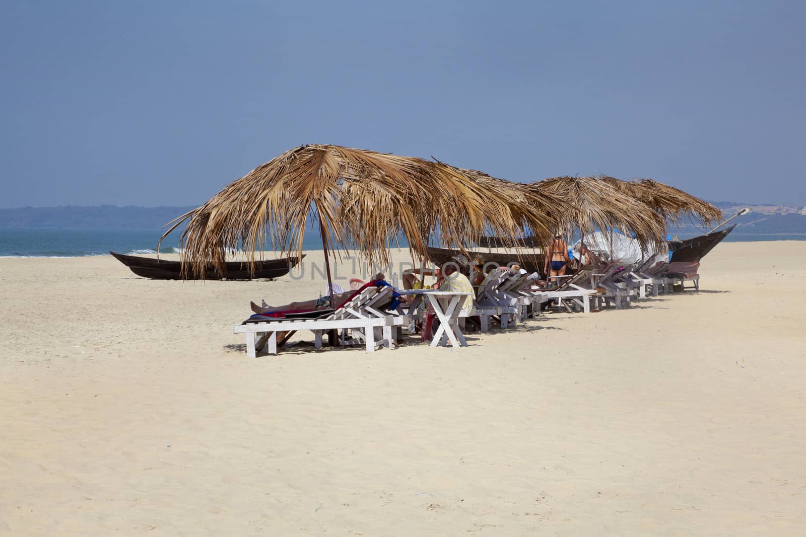 Landscape of secluded beach with day trippers and holidaymakers basking in the sun under the shade of coconut parasols. White sand, blue sky, sea, surf and fishing boats for a backdrop. Generic tropica shot location, Goa India