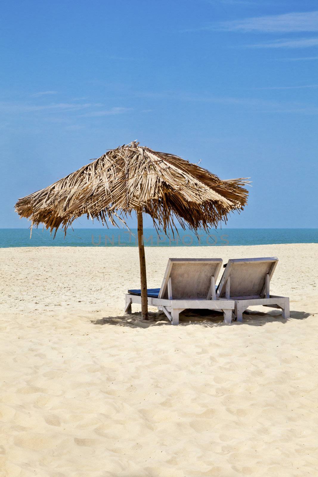 A generic vertical portrait of a tropical deserted beach with loungers under a coconut leaf parasol. Blue sky, ocean, gentle surf sound and clean sandy beach. Location, Goa India