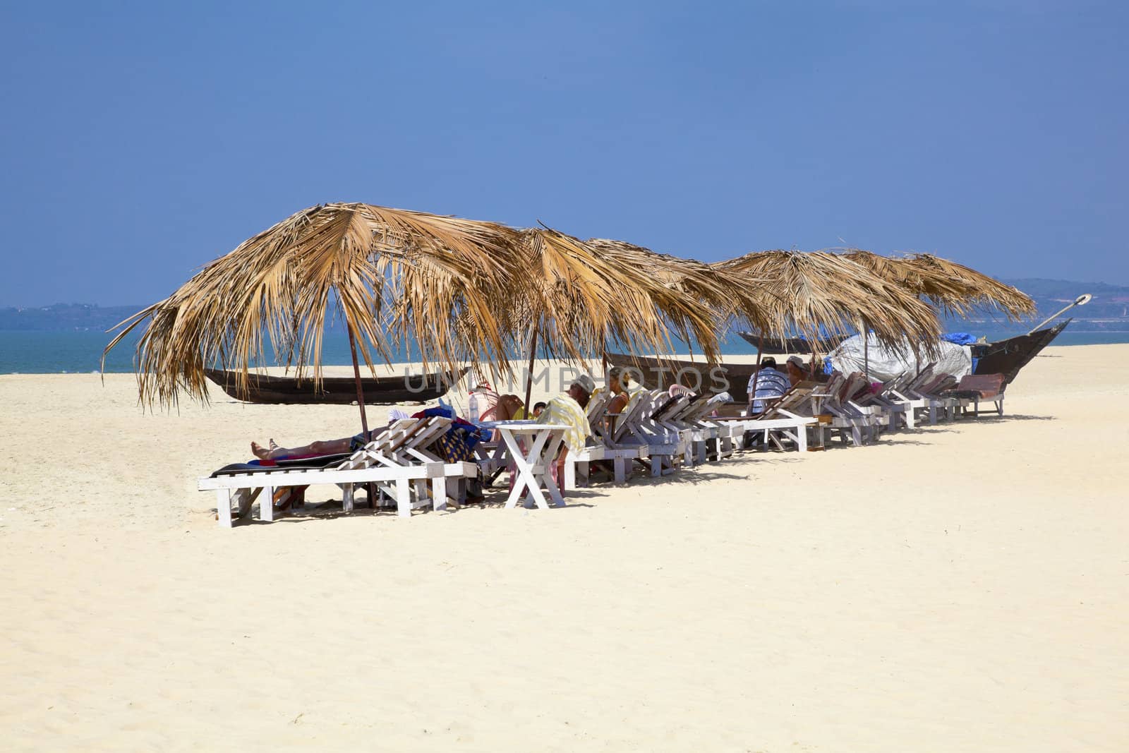 Landscape of secluded beach and a row of tropical parasols with day trippers and holidaymakers basking in the sun under the shade. White sand, blue sky, sea, surf and fishing boats for a backdrop. Generic tropical shot location, Goa India