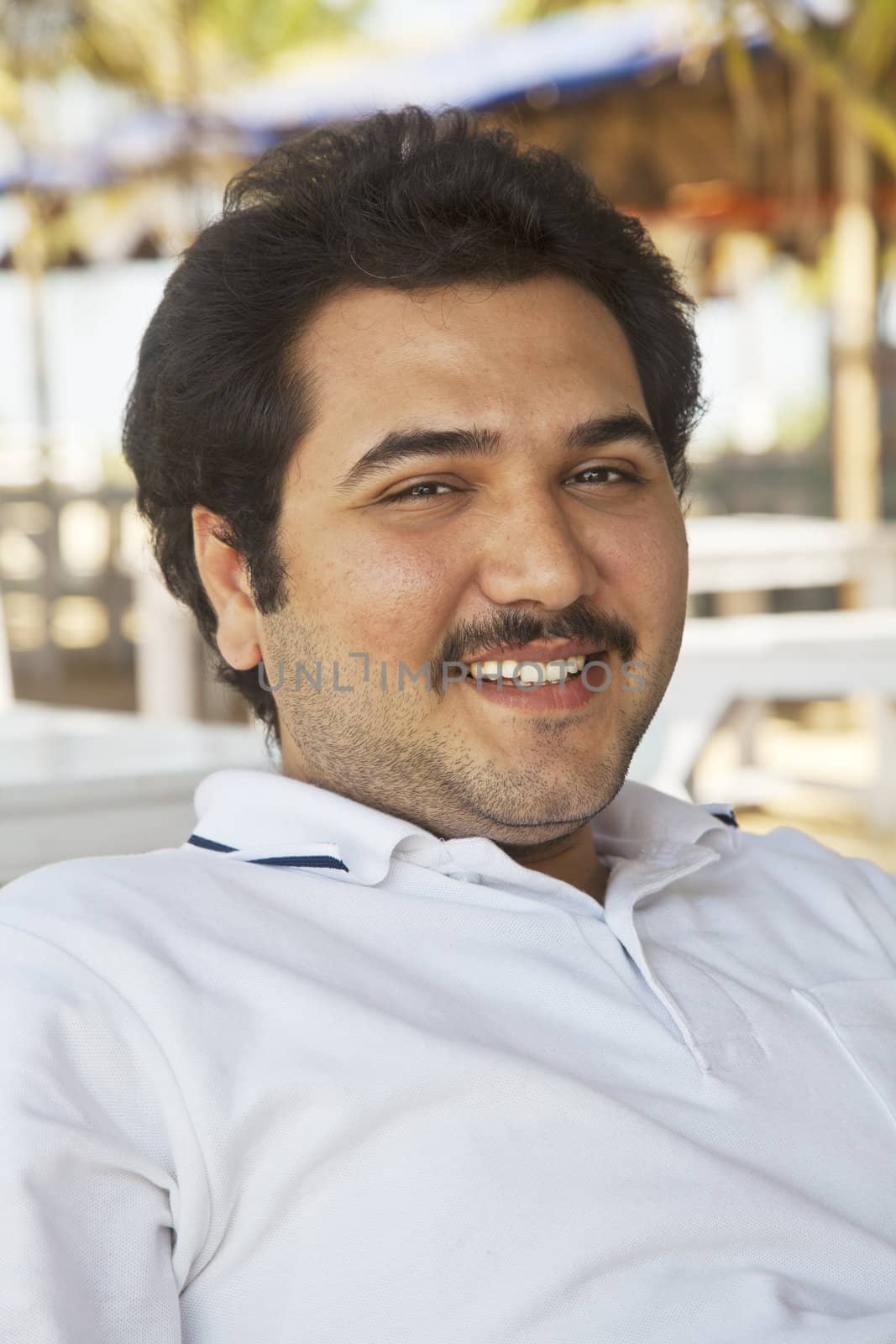 vertical portrait of a smiling young adult male, combed back hair, mustache, unshaved, celebrity look a like, showing teeth, wearing white pole shirt, in the shade outdoors leaning back in a relaxed posture