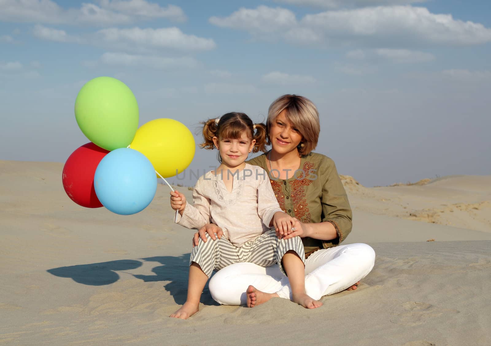 happy mother and daughter with colorful balloons sitting on sand by goce