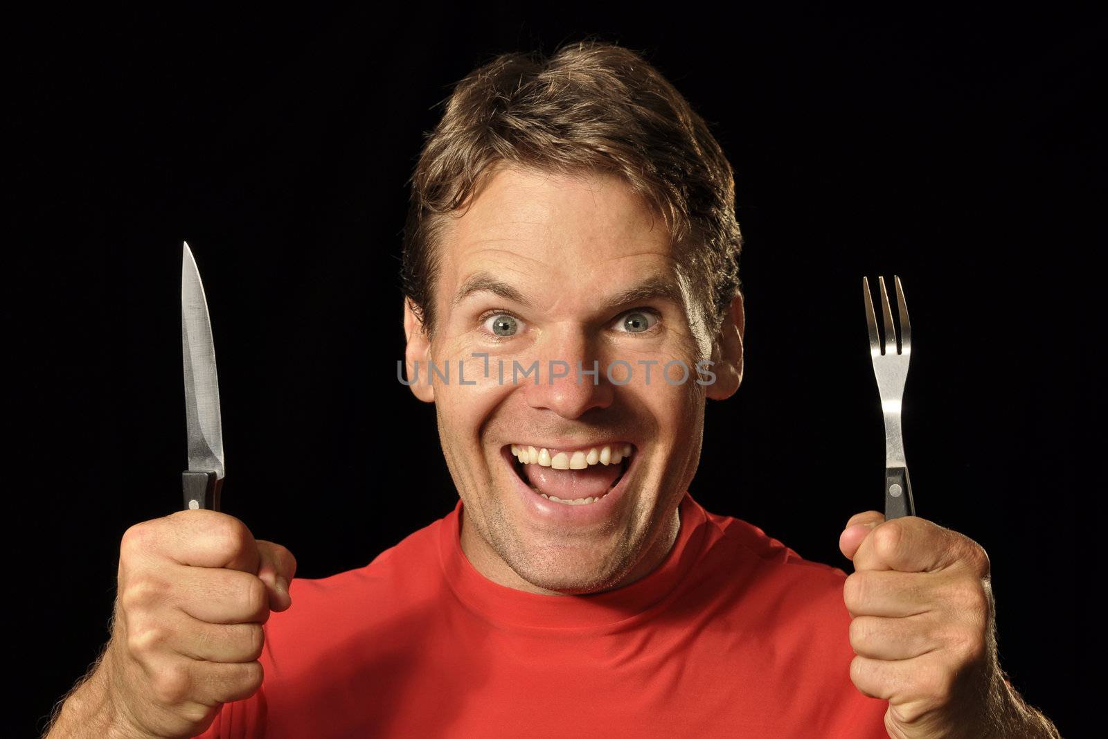Handsome hungry Caucasian man holds knife and fork while making excited face on black background