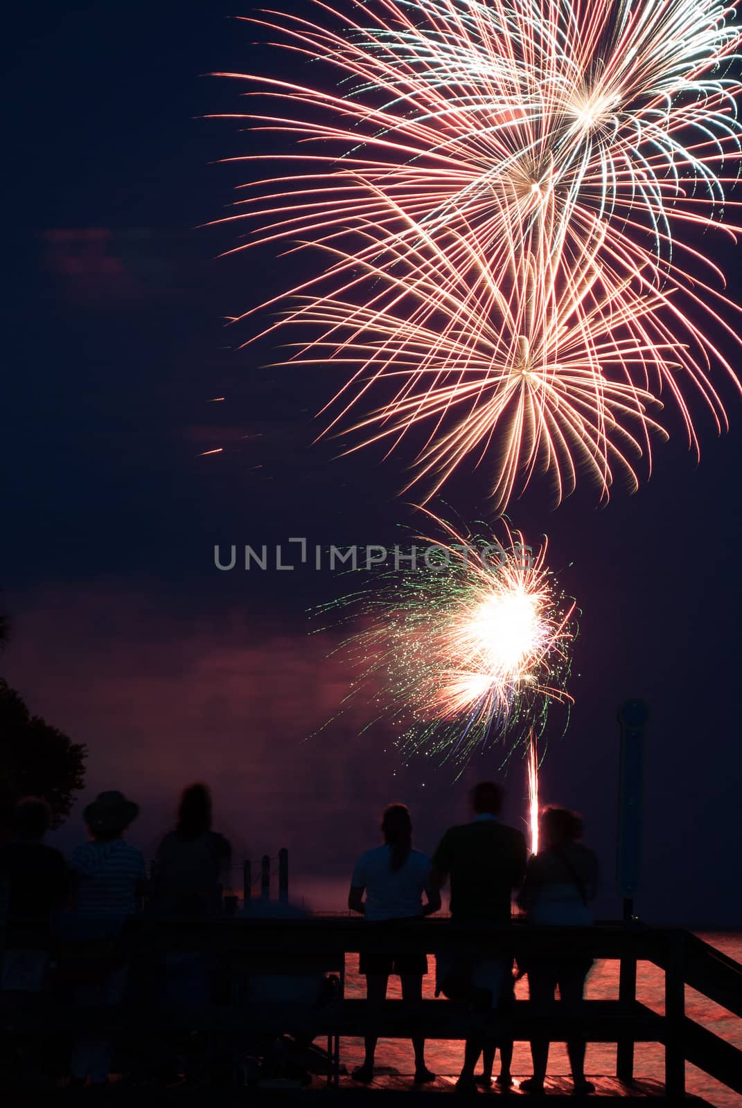 Photograph of various firework bursts from a 4th of July Celebration on Saint Simons Island Georgia.  Fireworks are shot off over water and reflection backlights people in foreground.