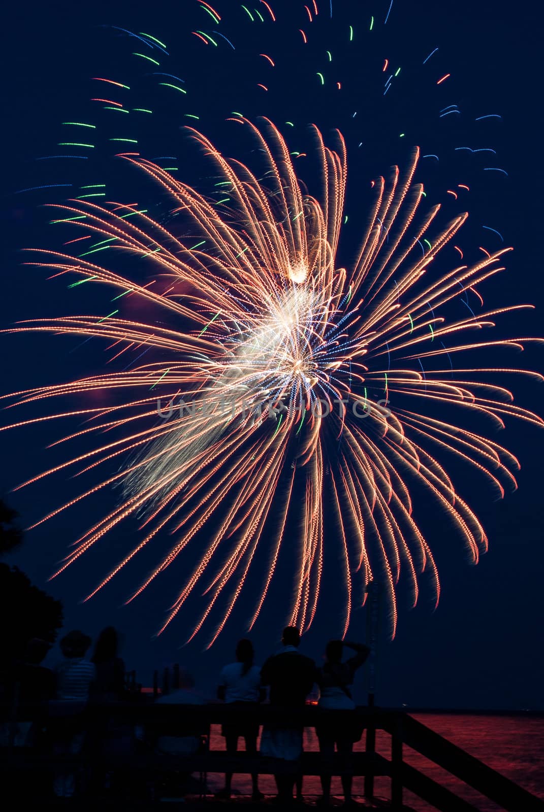 Photograph of various firework bursts from a 4th of July Celebration on Saint Simons Island Georgia.  Fireworks are shot off over water and reflection backlights people in foreground.