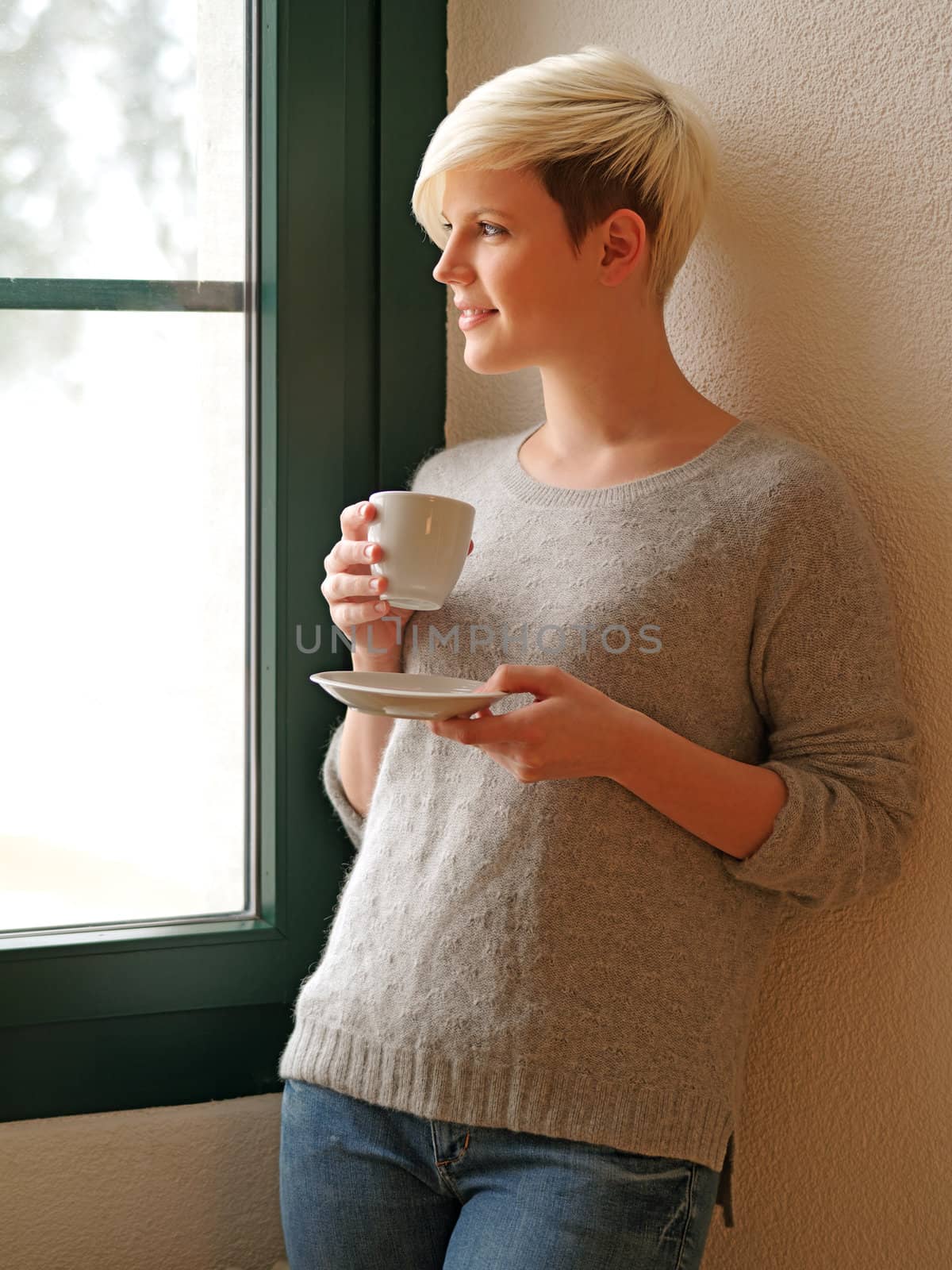 Photo of a beautiful young female drinking from a cup and looking out the window during winter.