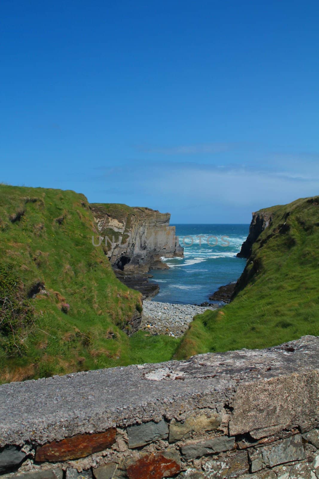 Small bay and grassy cliffs with stone fence separating the beach from the road in Ireland
