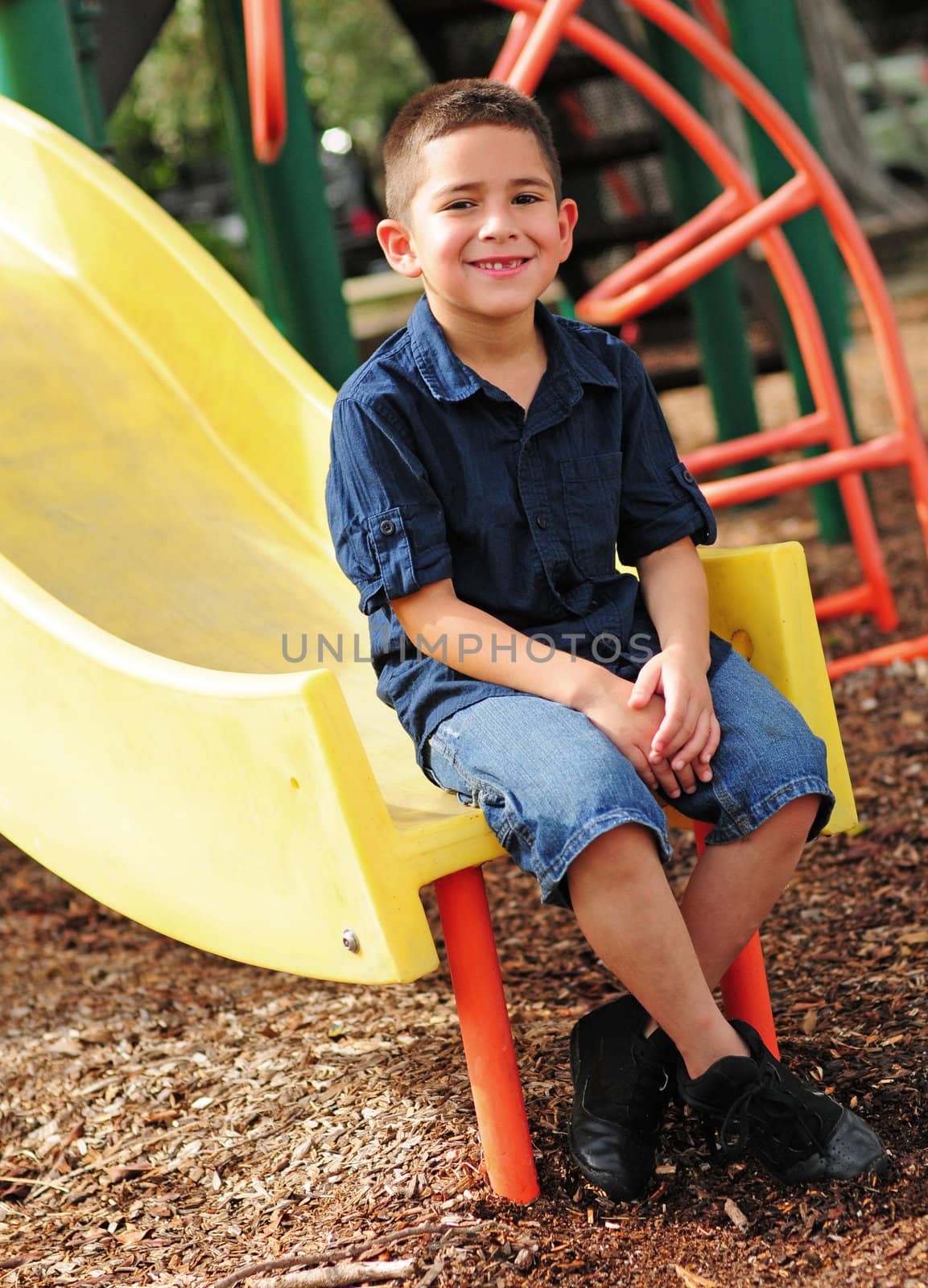 Happy young boy sitting at bottom of slide and having fun at outdoor park