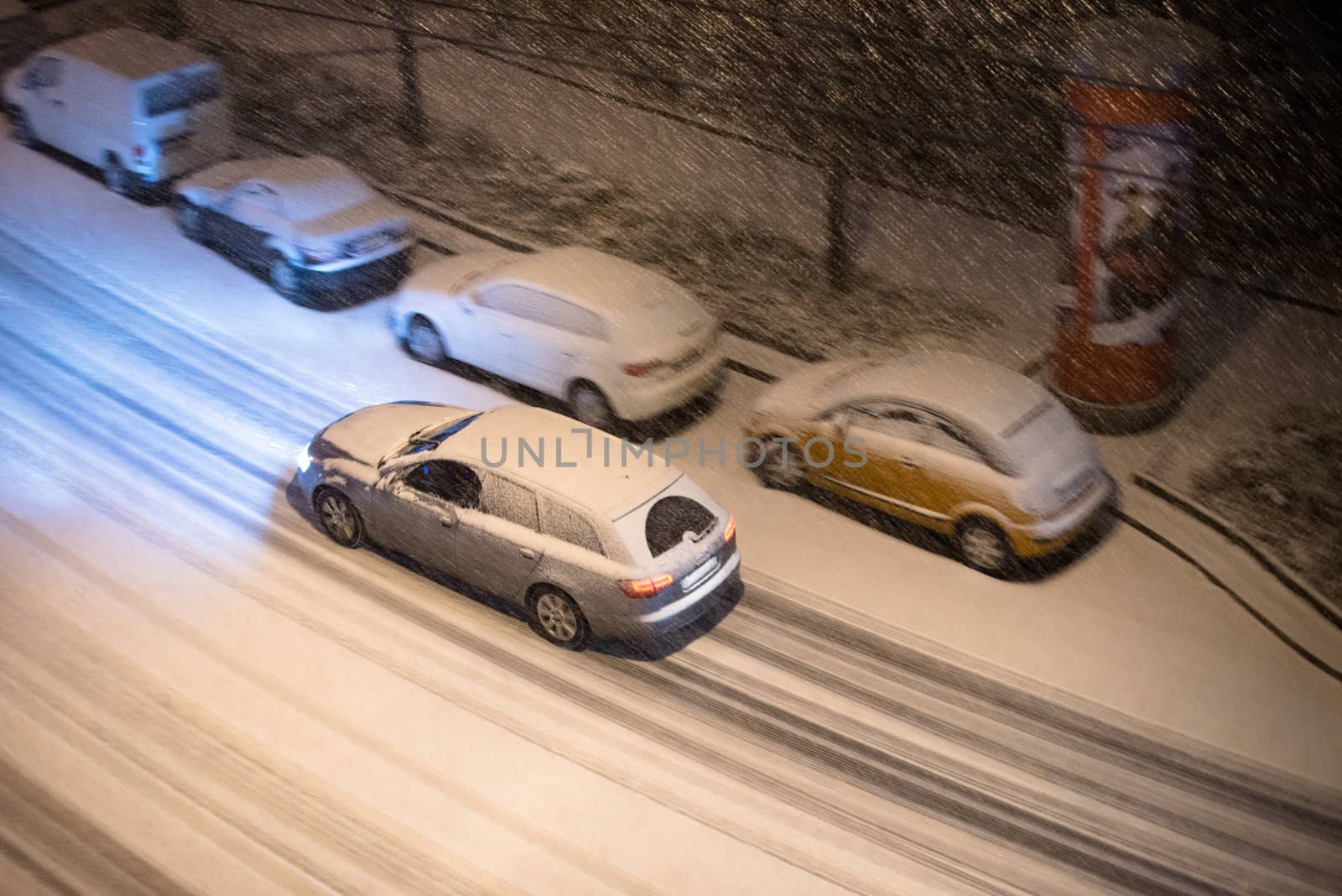 View from above on cars parked along a street and one car driving at night in winter snow