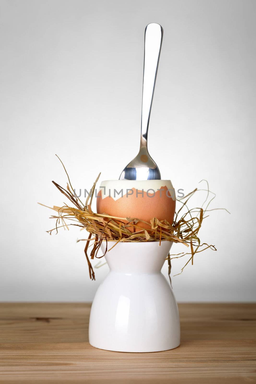 Easter egg in hay nest on white stand. Composition with egg ready to eat and spoon on wooden table background. Front view