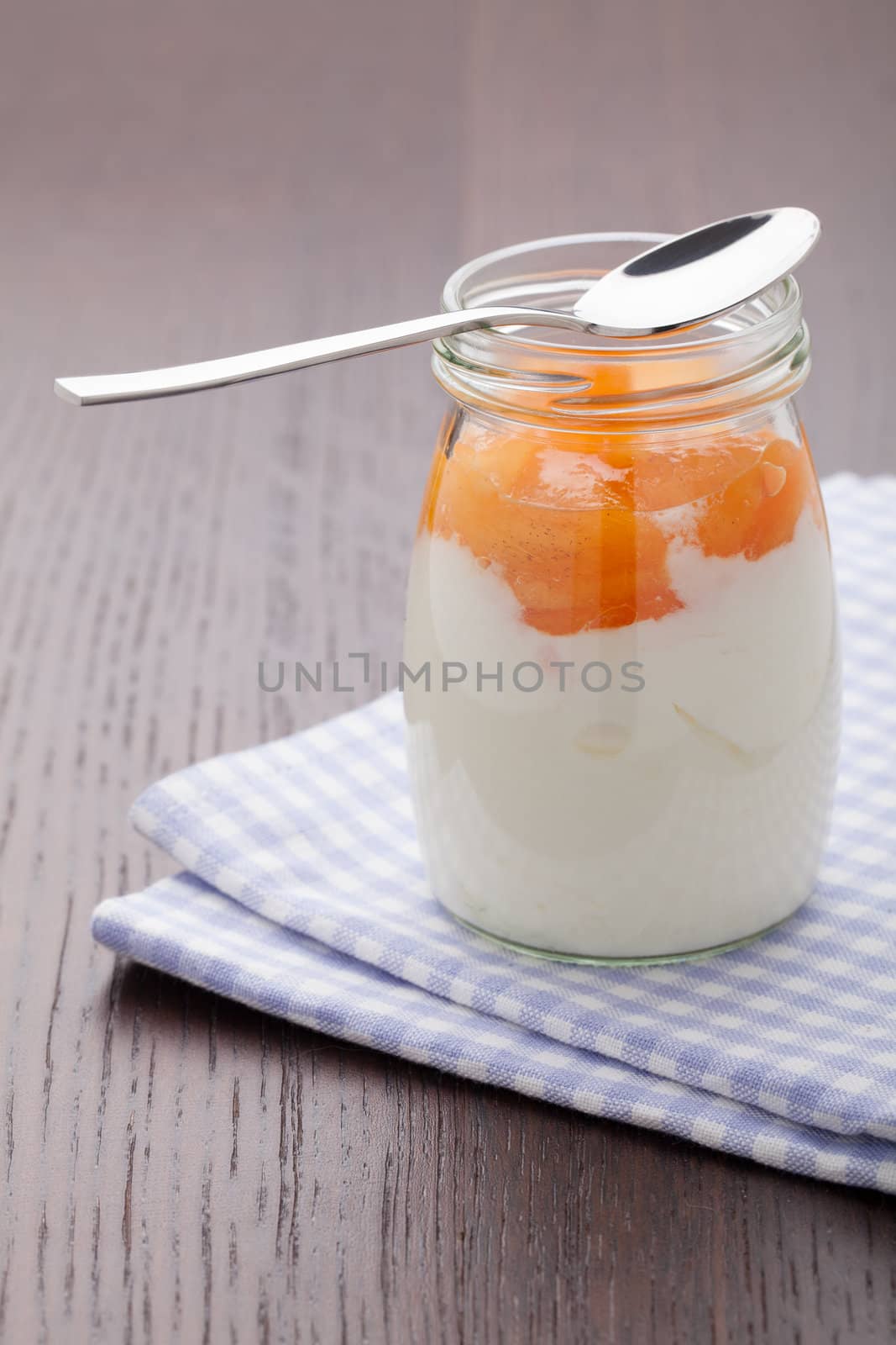 Homemade milk yogurt with peach or apricot jam in glass pot and metal spoon served on linen napkin, wooden table